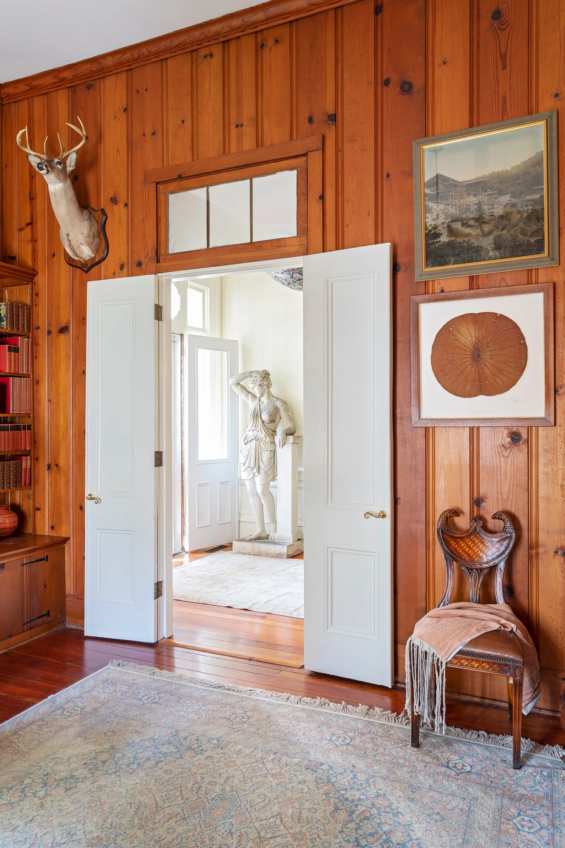A wood-paneled room features open white double doors leading to a brightly lit hallway containing a classical stone statue.