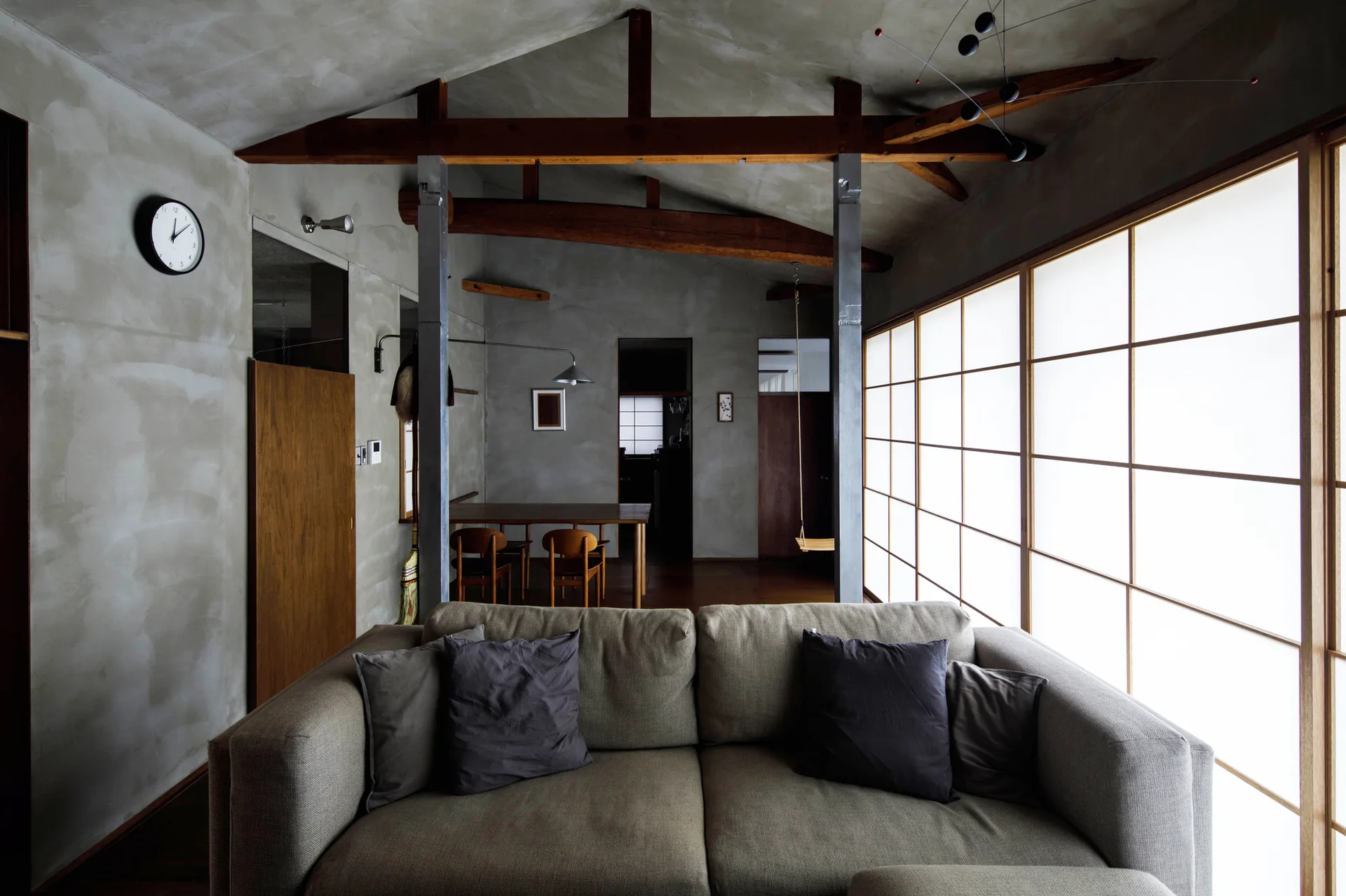 A minimalist living room features a grey fabric sofa, exposed wooden ceiling beams, textured plaster walls, and large shoji paper screens.
