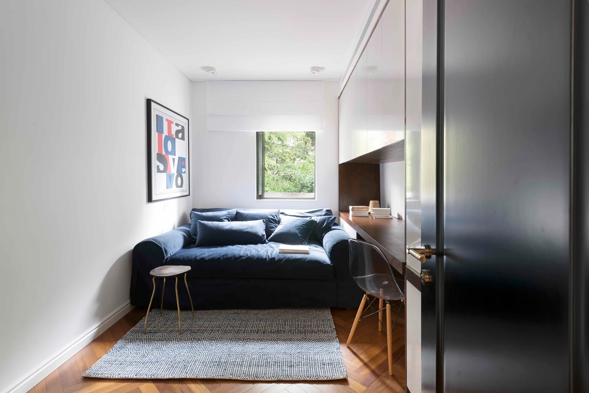 A small home office featuring a deep blue sofa under a window, an integrated wooden desk with white glossy cabinets, and herringbone parquet floors.
