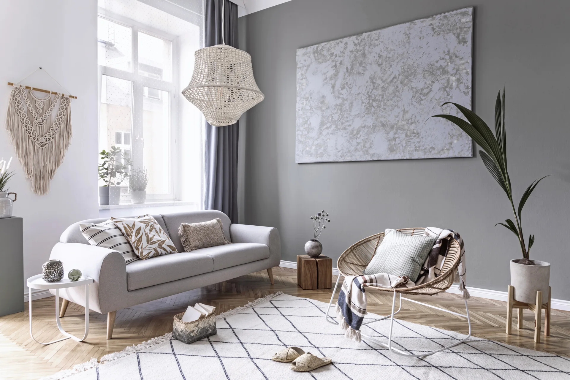 A grey sofa and a rattan rocking chair sit on a grid-patterned rug in a living room with sage green walls and herringbone wood floors.