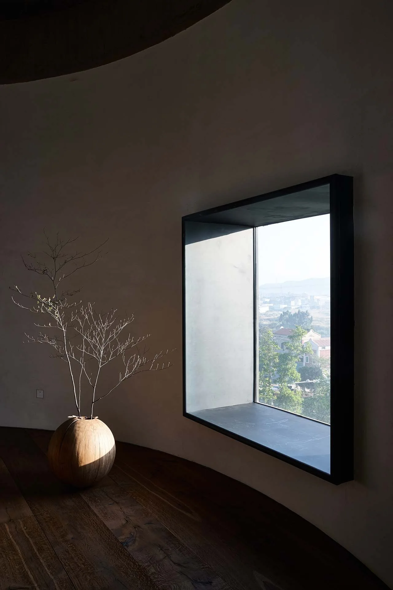 A large rectangular inset window set into a curved white plaster wall next to a spherical wooden vase with a bare branch on a wood floor.