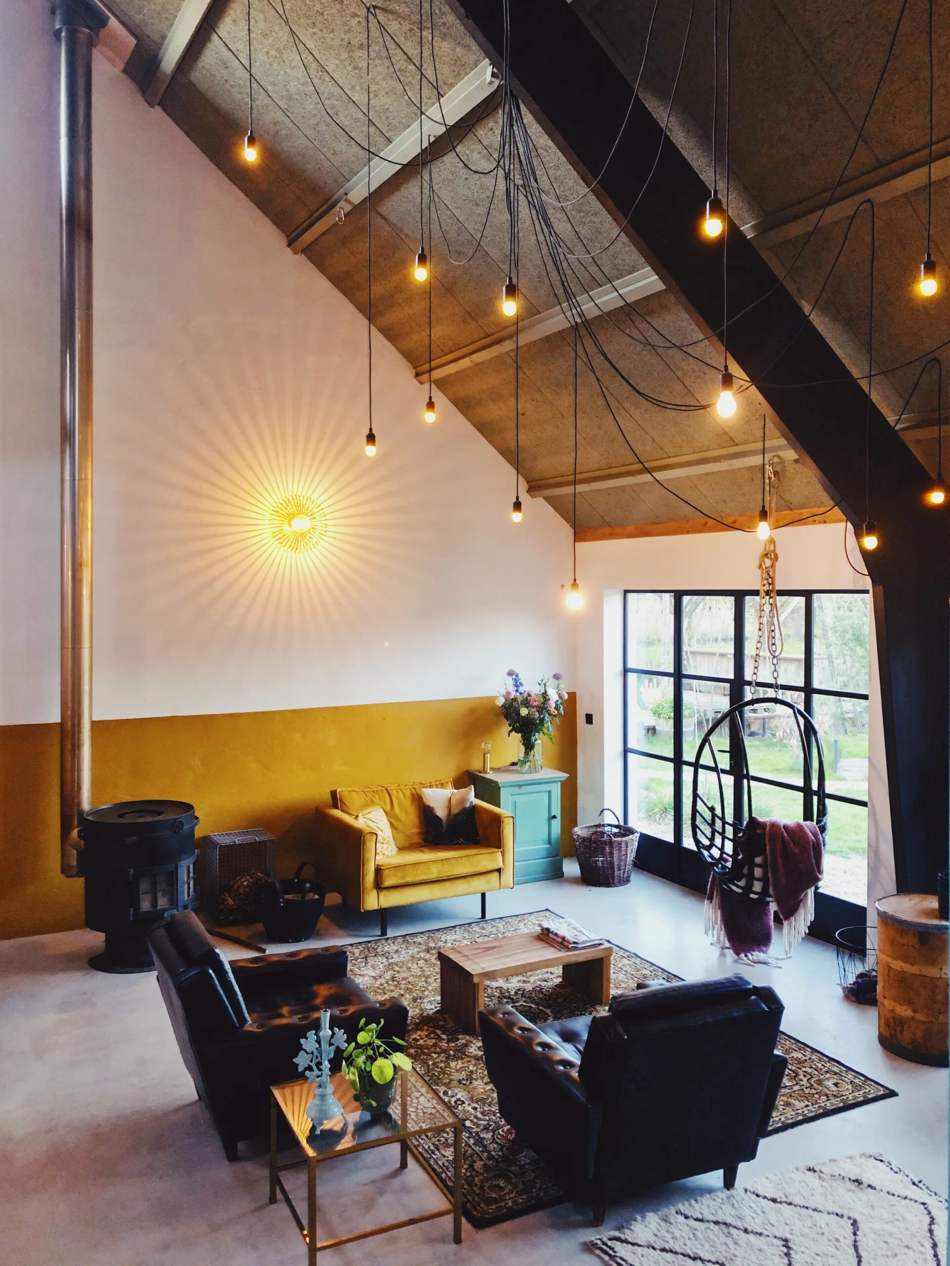 A spacious living room with vaulted ceilings featuring cluster pendant lights, a mustard yellow accent wall, two black leather armchairs, and a hanging chair.