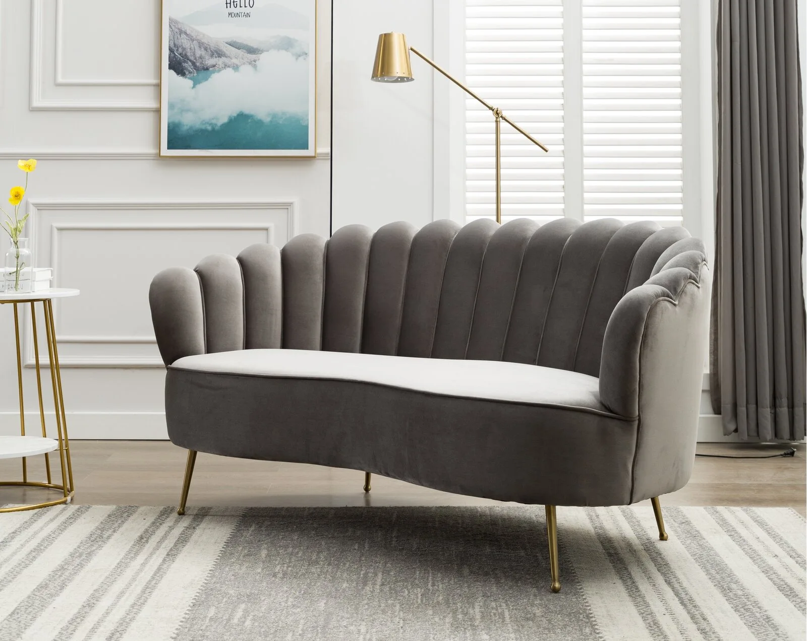 A grey curved velvet sofa with channel tufting and gold legs sits on a striped rug in front of a white wall with decorative molding.