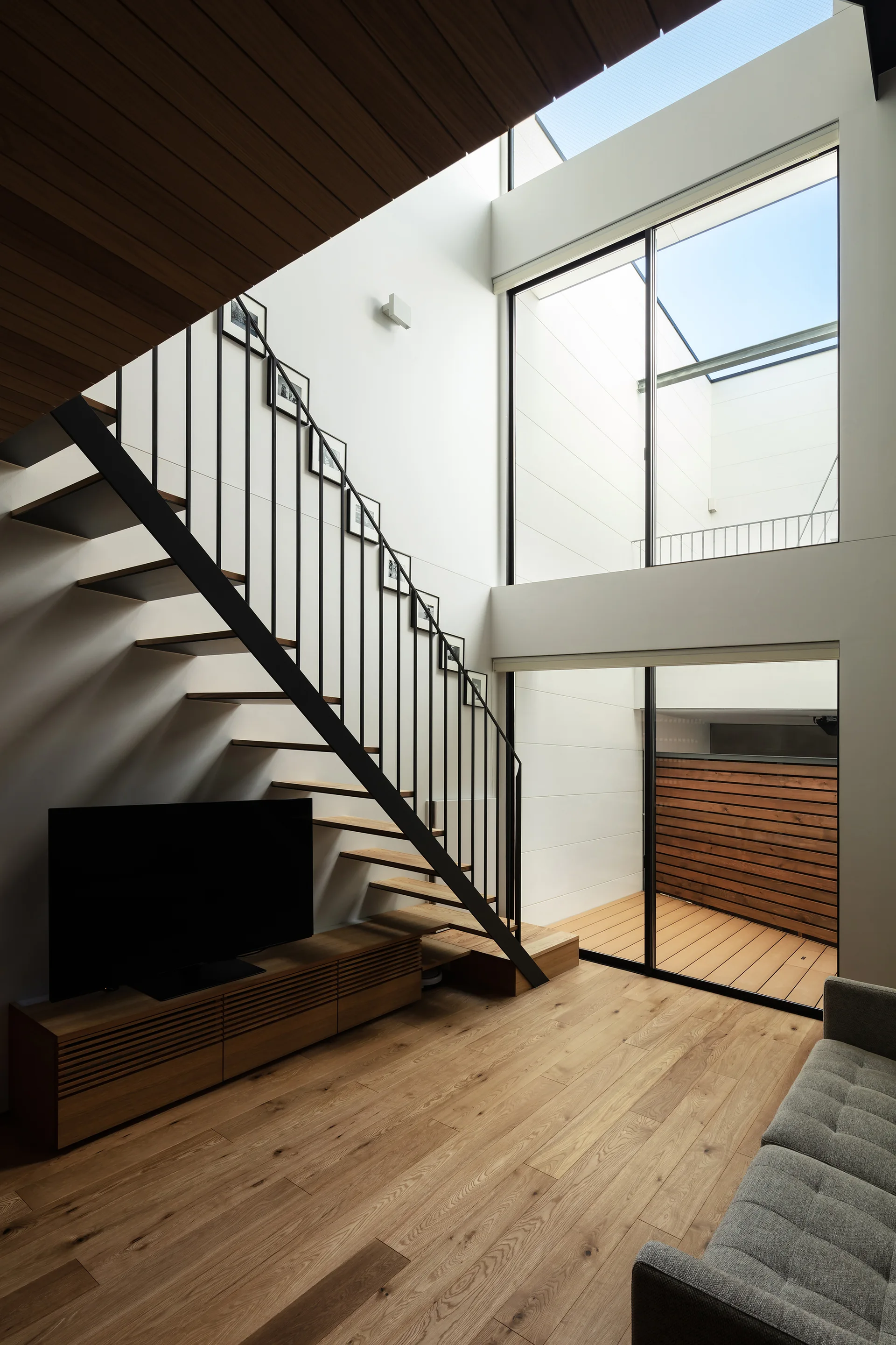 A modern living room featuring a wooden open-riser staircase, a low wooden media console, a grey sofa, and a tall glass wall.