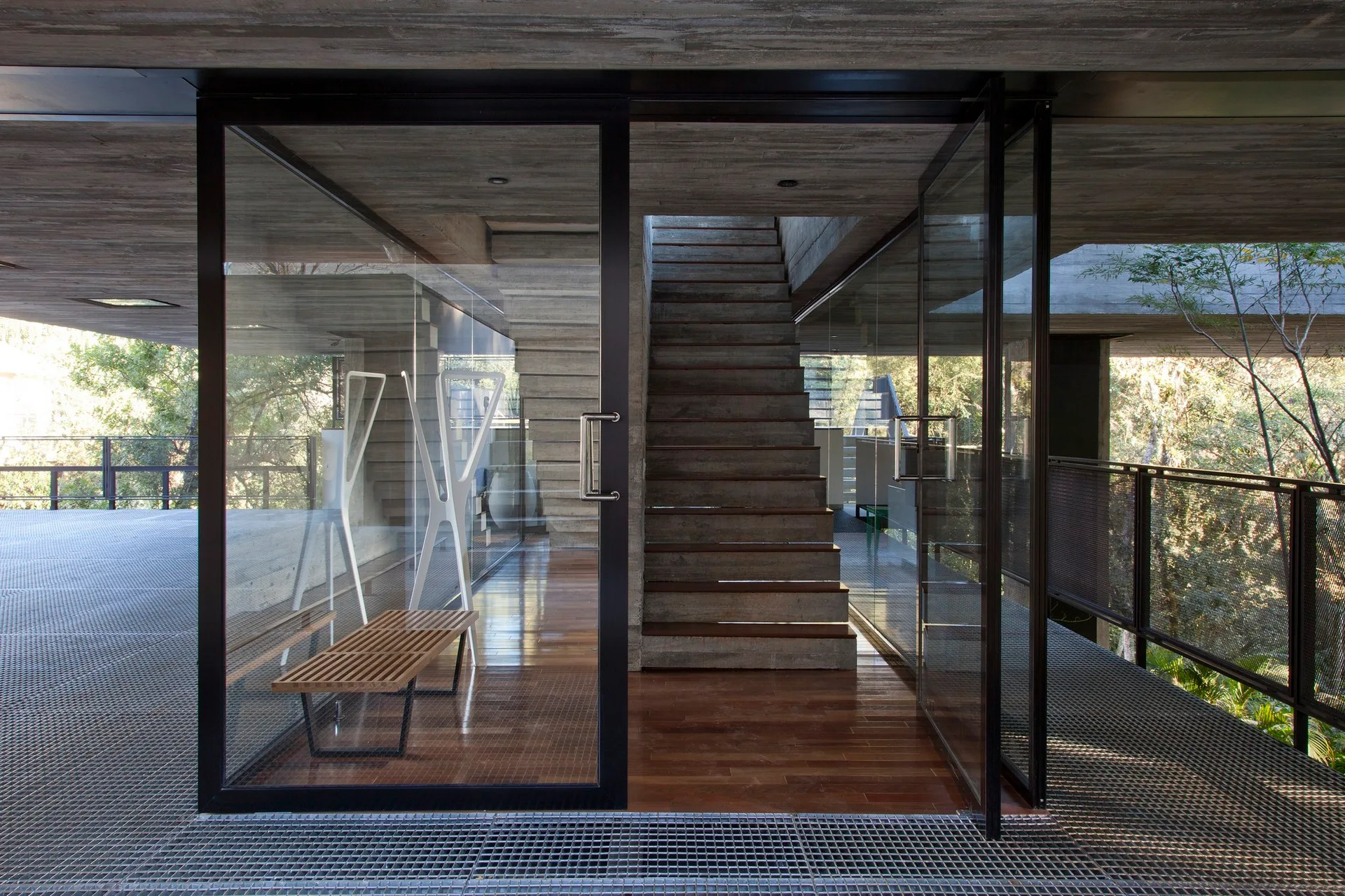 An entryway with metal-framed glass doors features a central concrete staircase, a slatted wood bench, and a transition from metal grating to wood flooring.