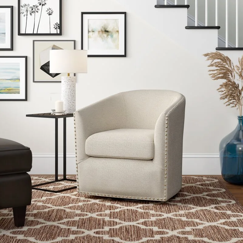 A cream upholstered barrel swivel chair with gold nailhead trim sits on a brown patterned rug next to a black side table and a staircase.
