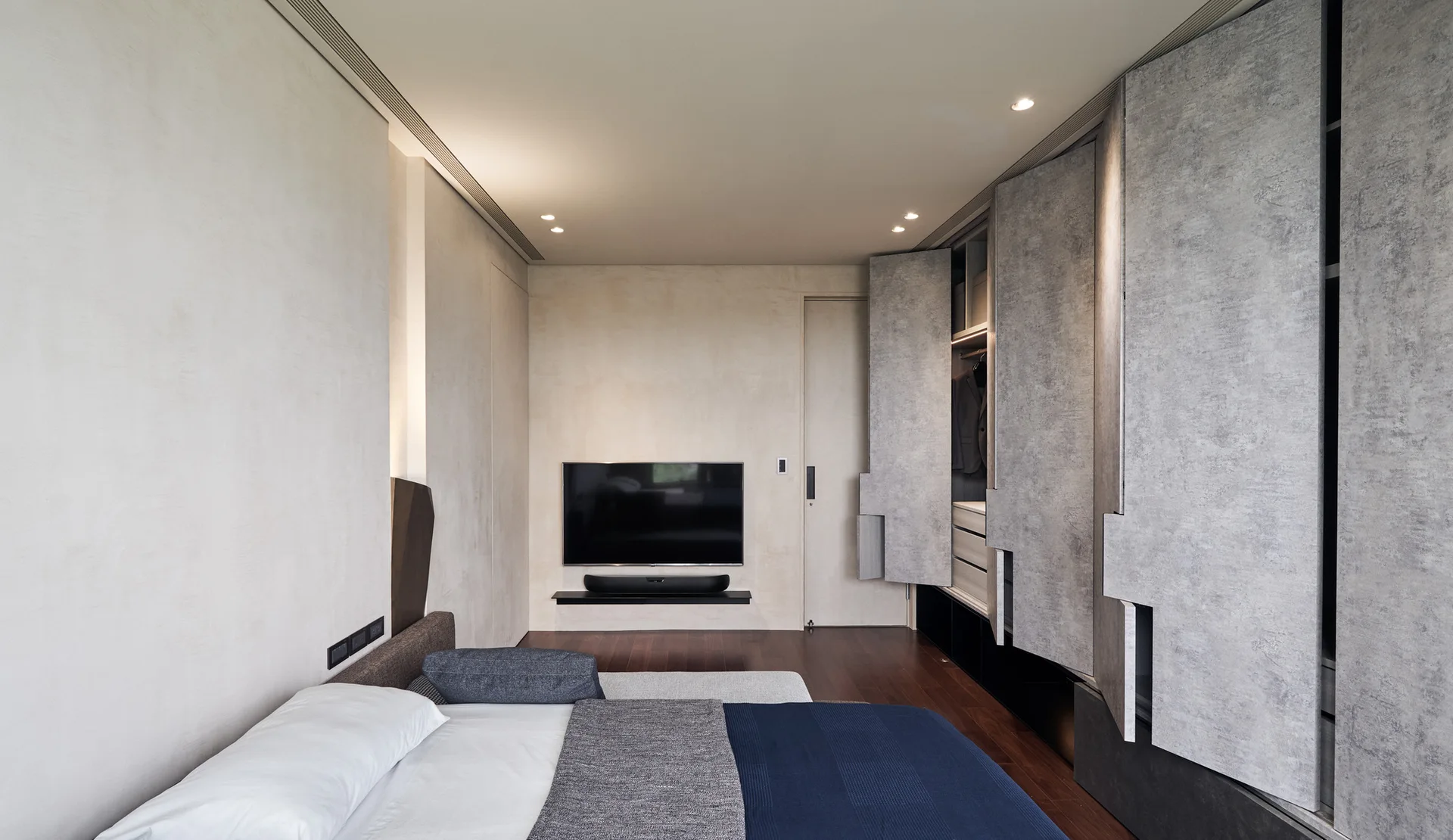 A minimalist bedroom features a bed with blue bedding, textured beige walls, a wall-mounted TV, and an integrated closet with grey stone-patterned doors.