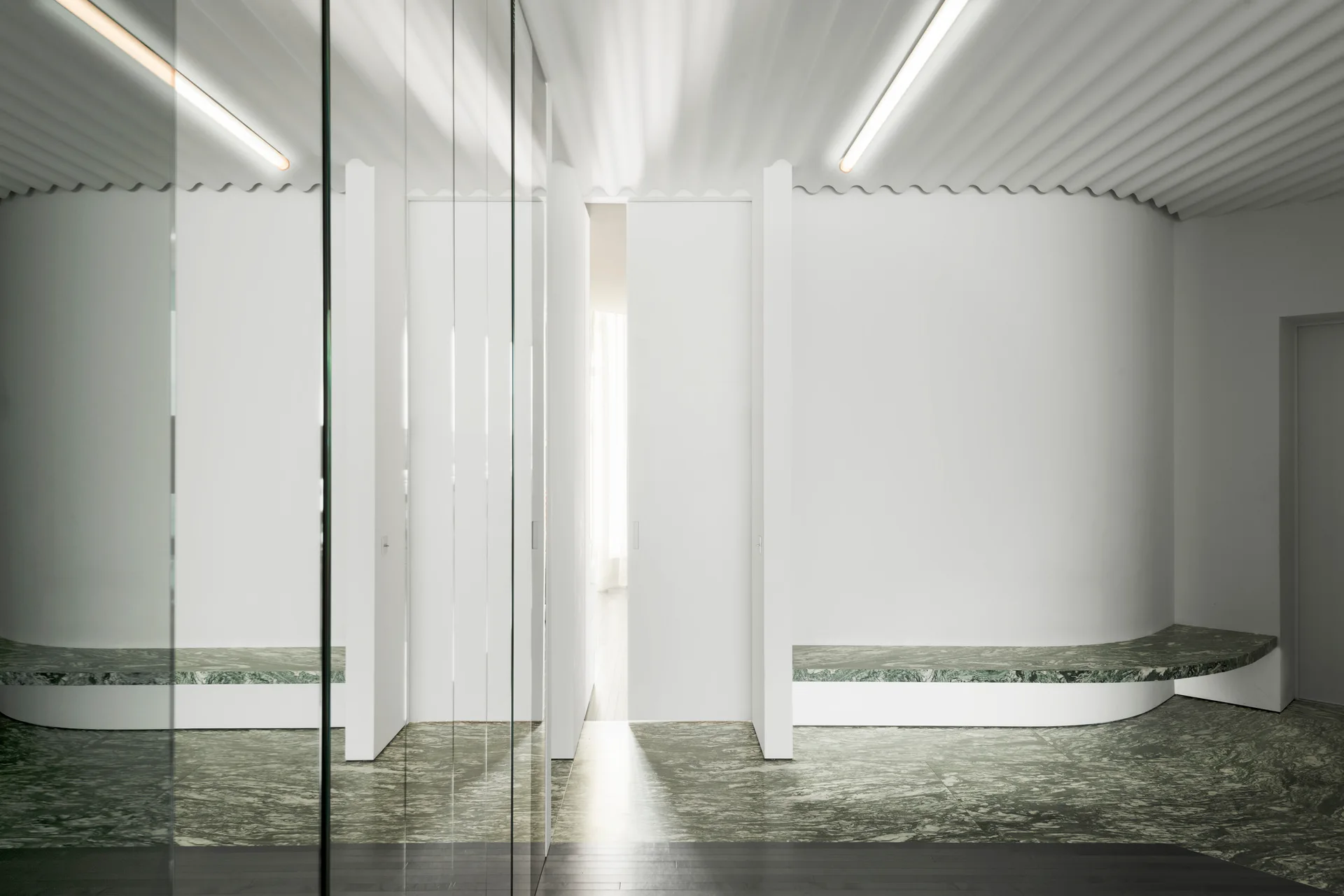 A minimalist white hallway features curved green marble flooring, matching built-in benches, a corrugated ceiling, and linear tube lighting.
