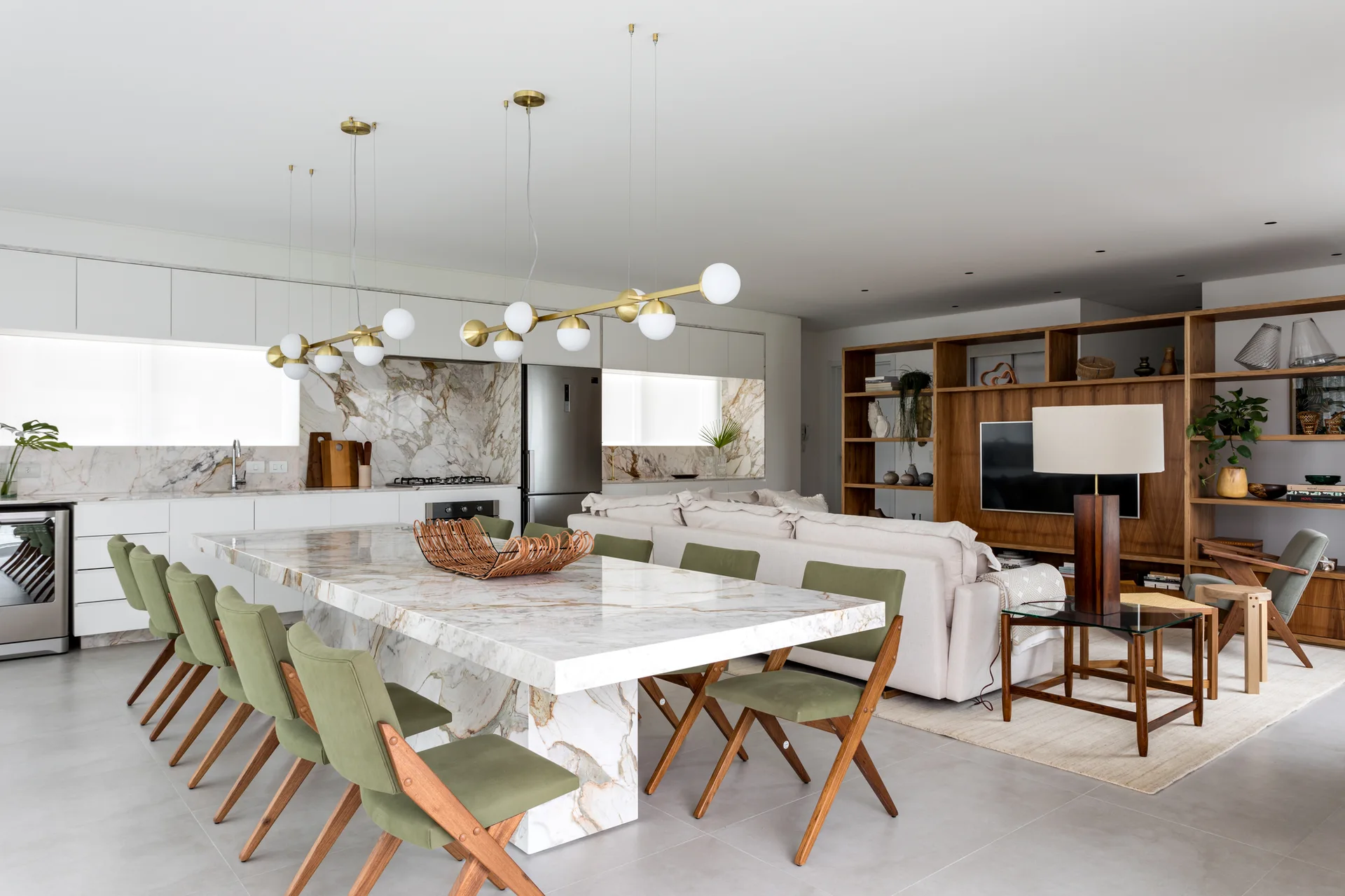 An open plan room featuring a large veined marble dining table with green chairs, a beige sectional sofa, and a custom wood shelving unit.
