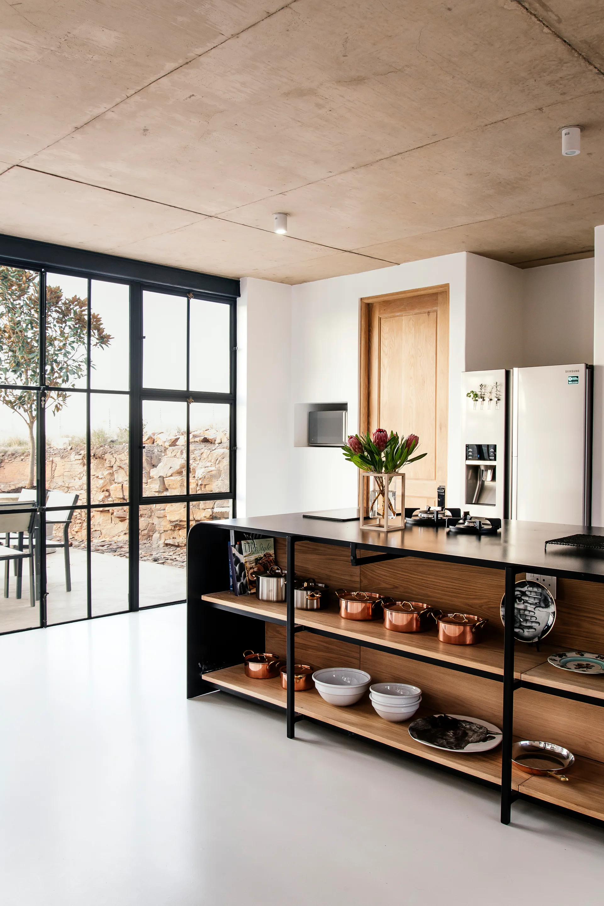 A modern kitchen featuring an open-shelved wood island with copper cookware, set against an exposed concrete ceiling and black metal-grid window.