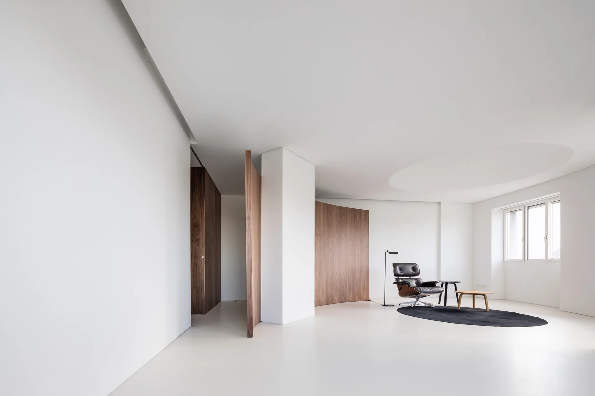 A minimalist white living room features a black leather lounge chair on a dark oval rug alongside curved wood veneer wall partitions.