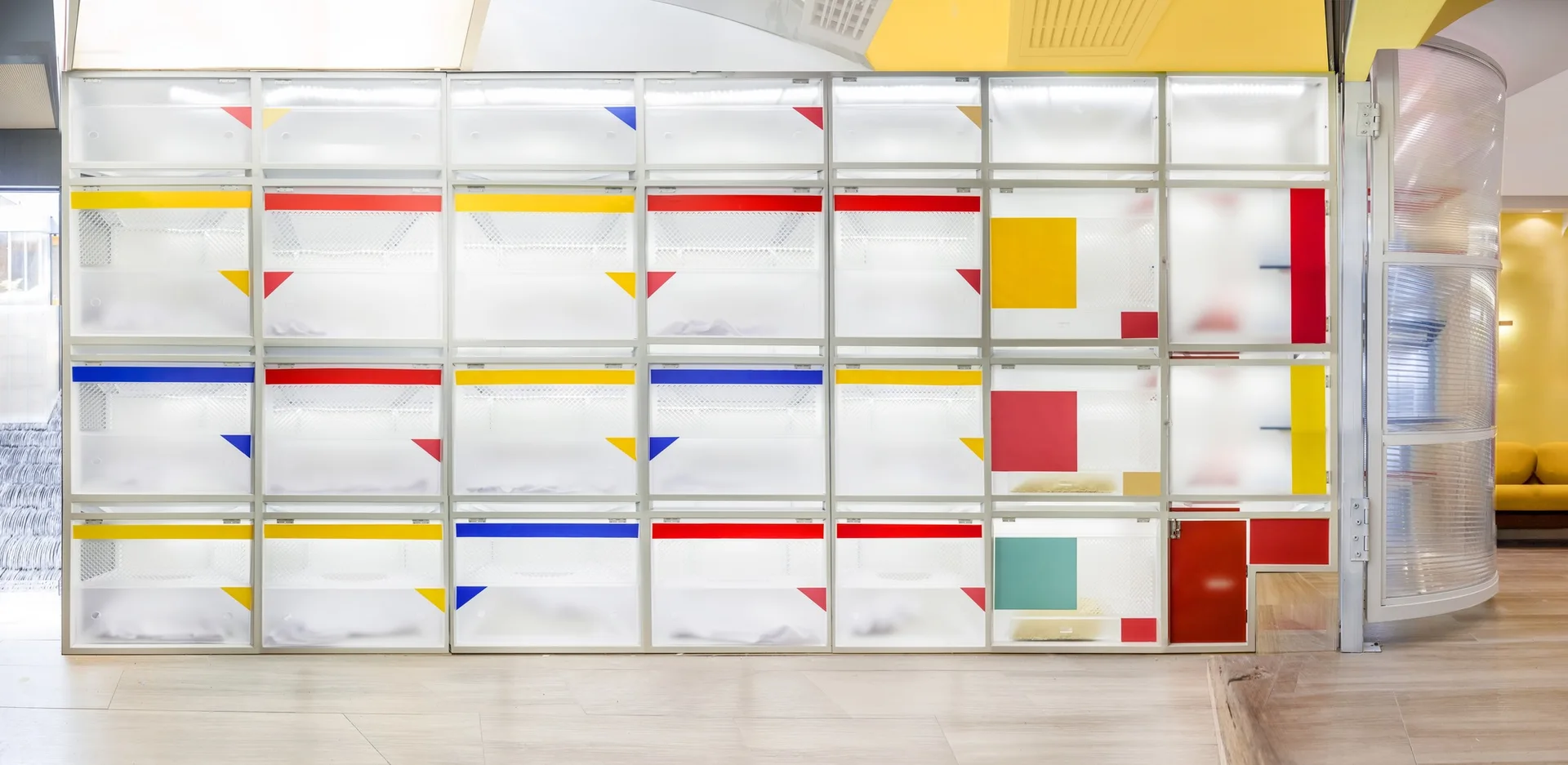 A wall-sized grid storage unit features translucent frosted panels accented with primary colored shapes and integrated interior lighting.
