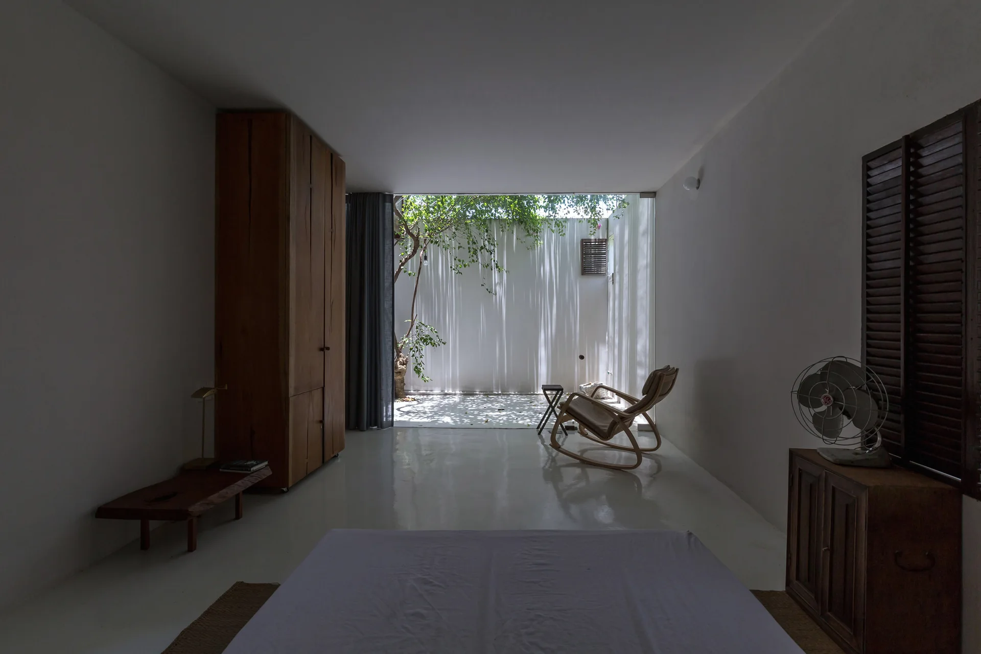 A minimalist white bedroom with a tall wooden wardrobe and a rocking chair overlooking a sunlit inner courtyard with a tree.