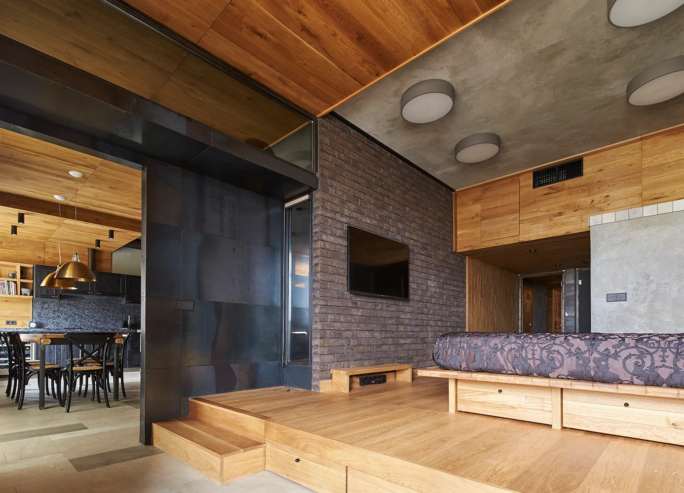 An industrial style bedroom features a raised wooden platform bed against a dark brick wall, with an adjoining kitchen in the background.