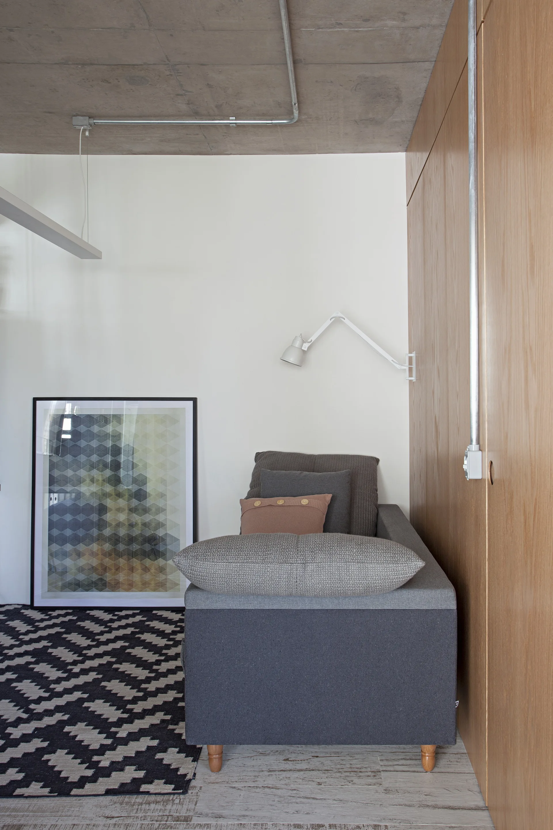 A gray daybed rests against a wood-paneled wall and white wall under a raw concrete ceiling with exposed conduit and a geometric rug.
