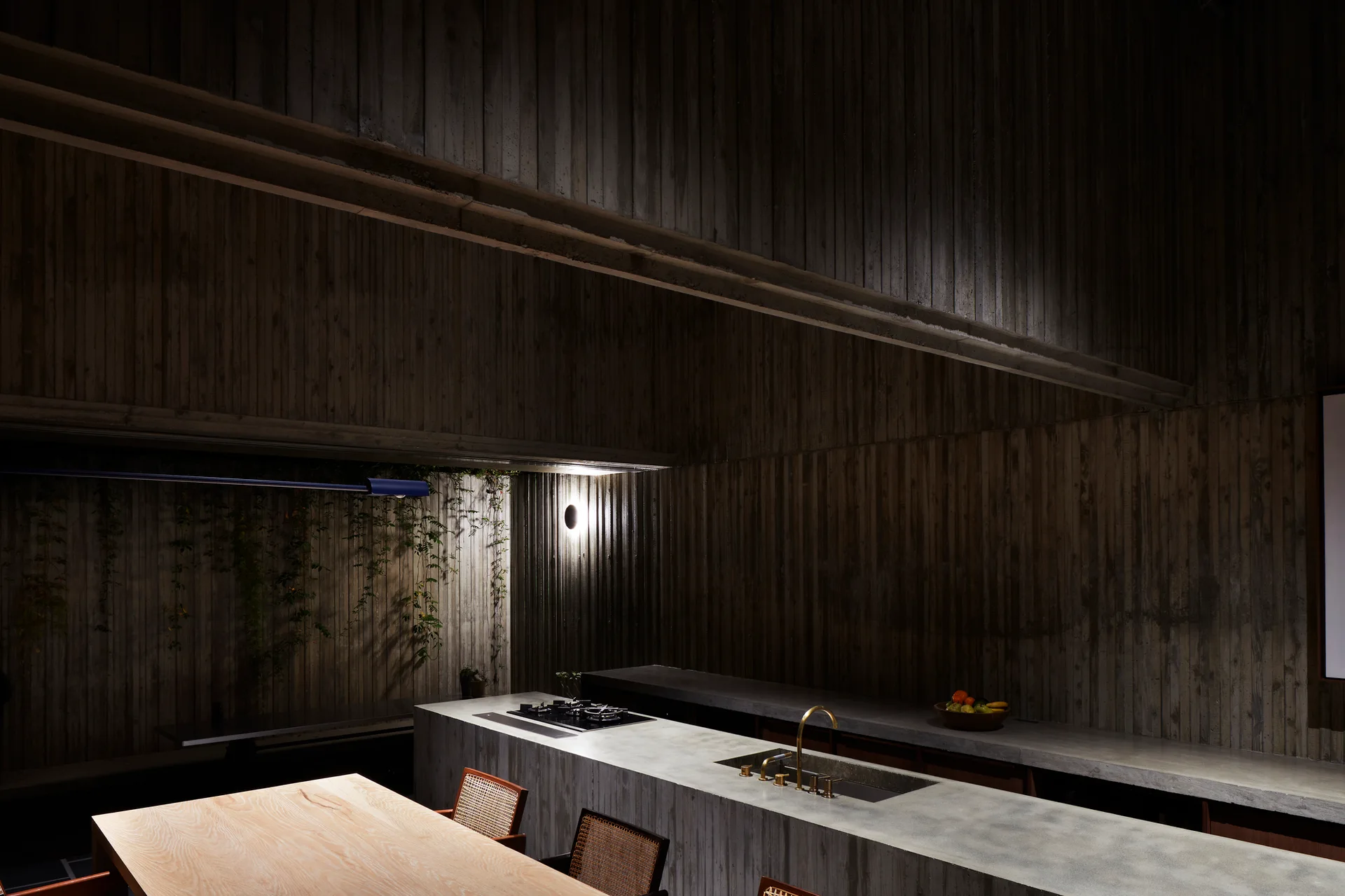A minimalist modern kitchen with a large concrete island, board-formed concrete walls, and a wooden dining table with rattan chairs.