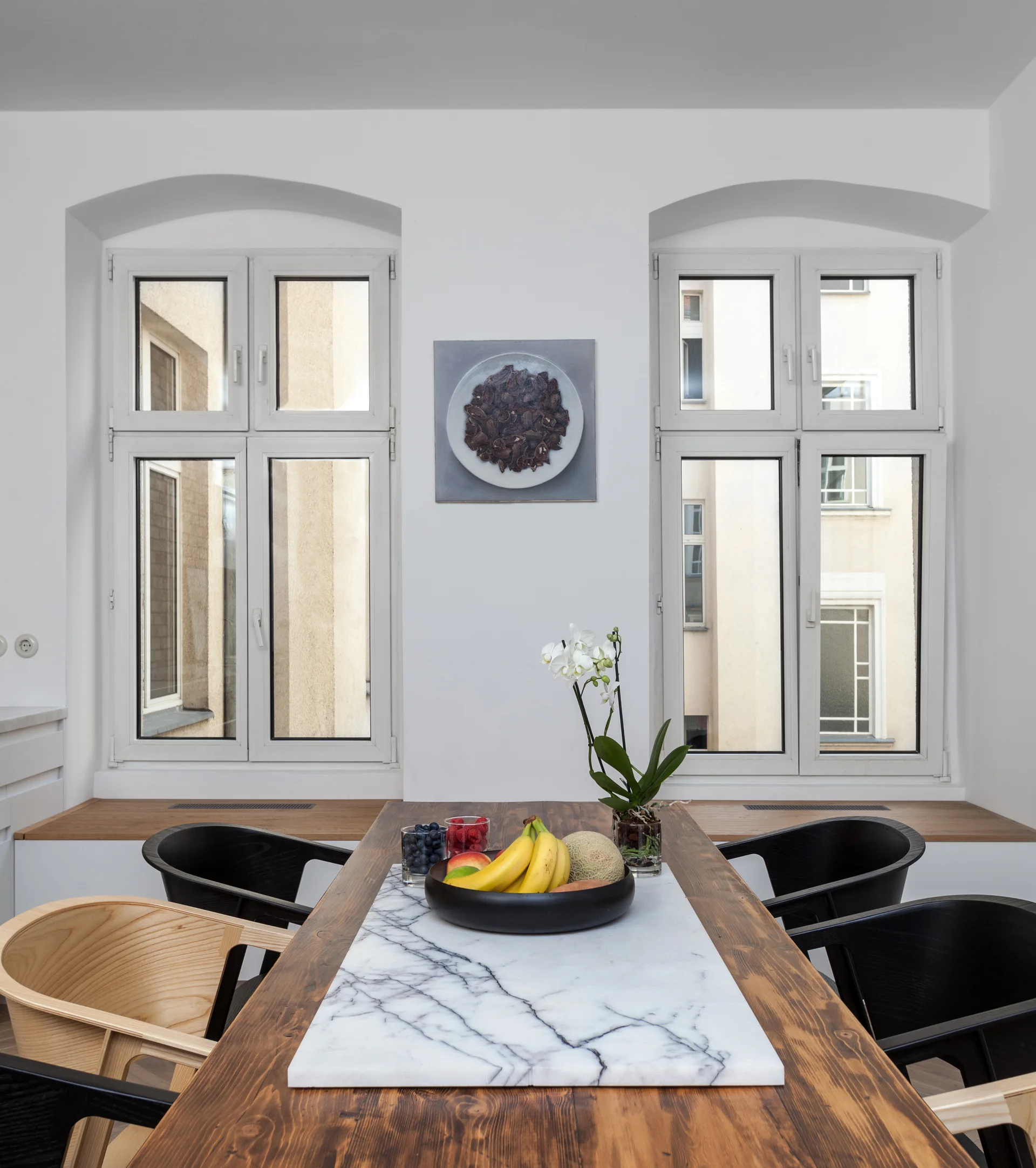 A wooden dining table with a white marble insert is surrounded by black and wood chairs in front of two arched windows and a wooden bench.
