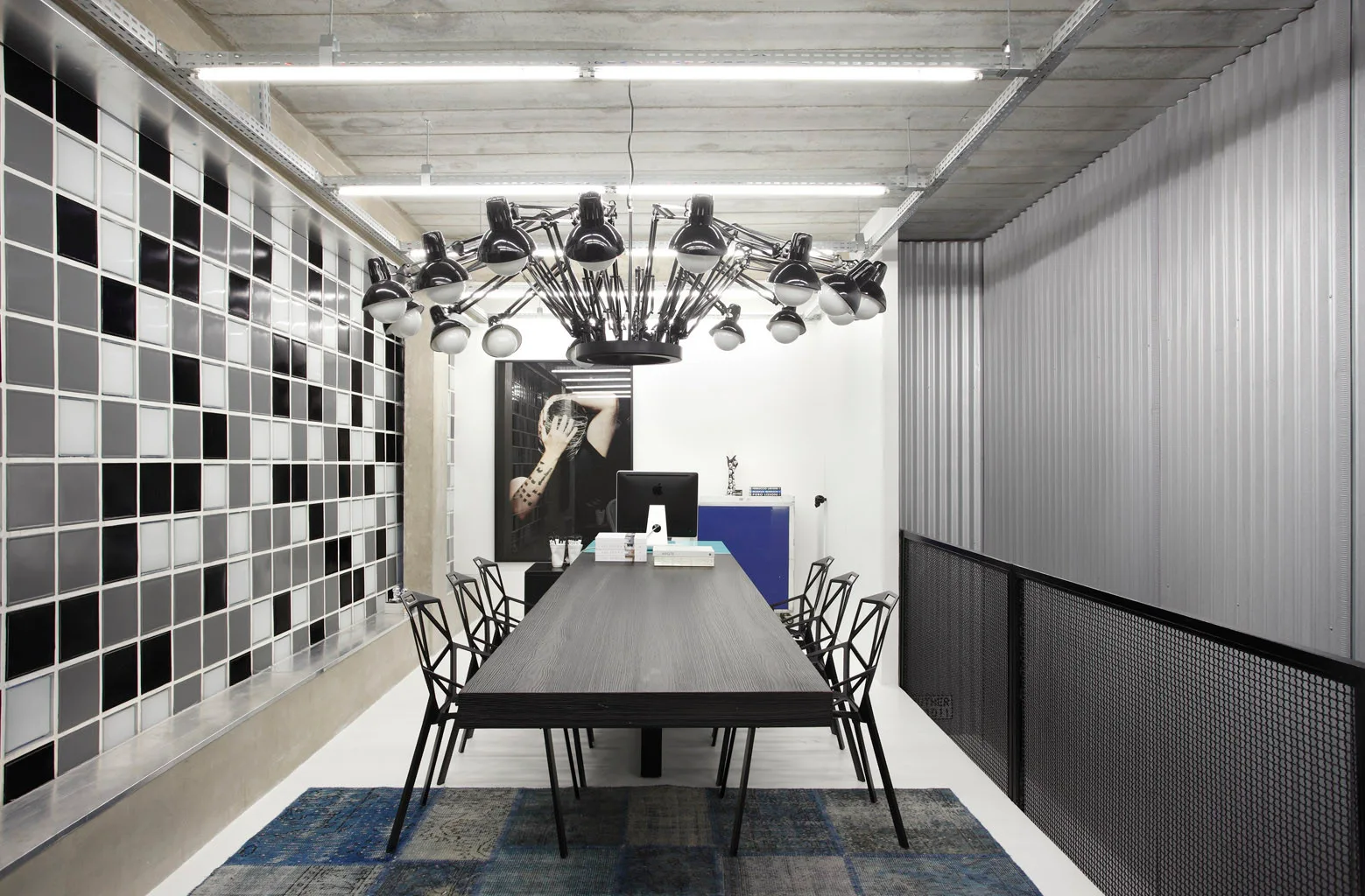An industrial workspace features a long black table, geometric metal chairs, a massive multi-arm black chandelier, and a black-and-white tiled wall.
