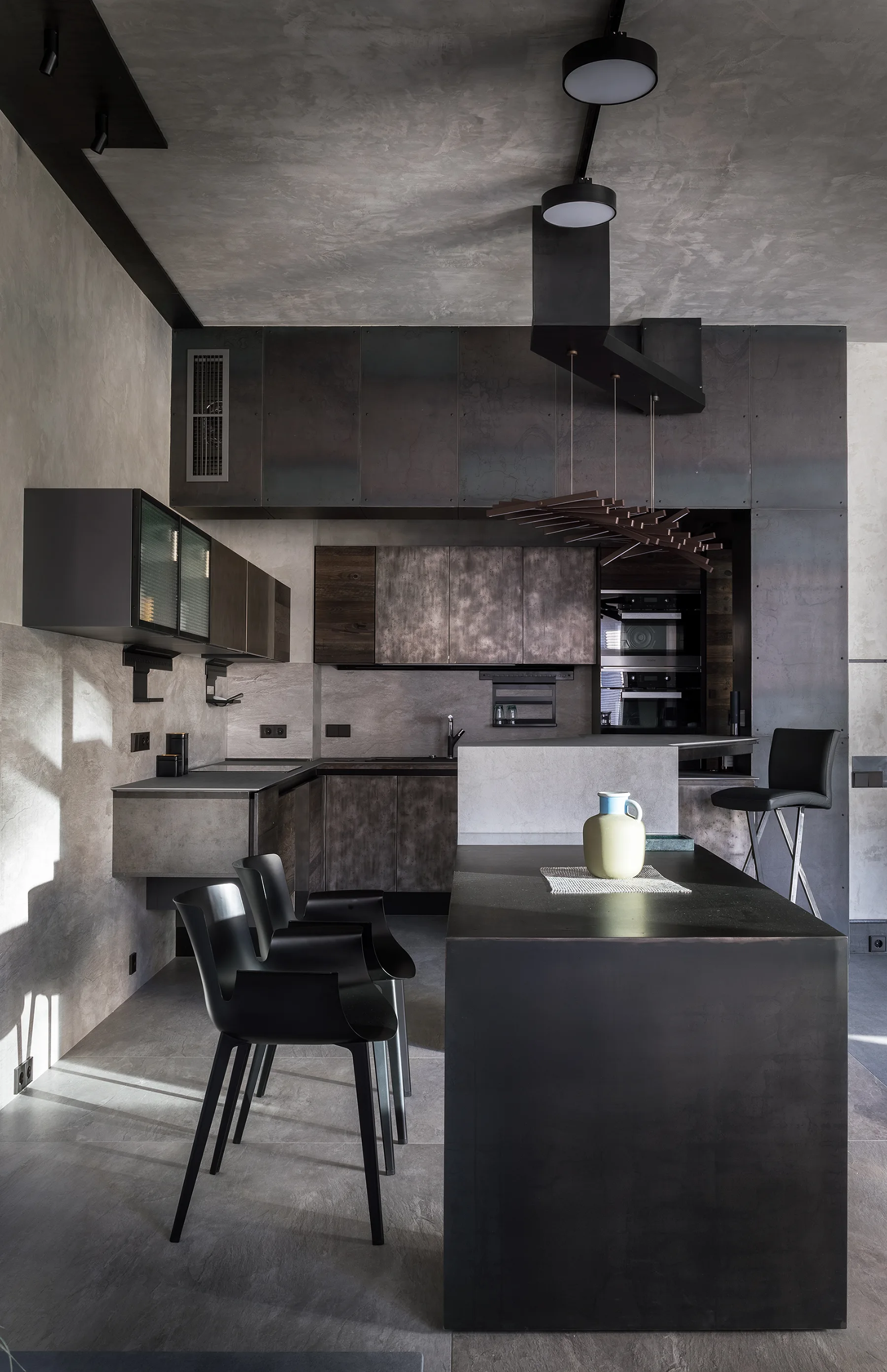 A dark industrial kitchen featuring textured gray concrete walls and ceiling, metallic dark grey cabinetry, black dining chairs, and a solid black island.