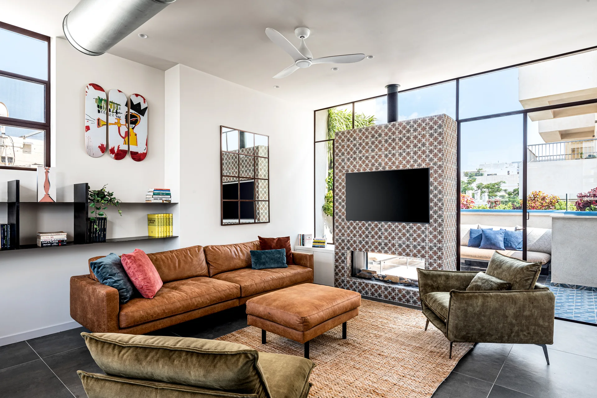 A contemporary living room featuring a brown leather sofa, olive velvet chairs, and a central fireplace tiled in a geometric pattern.