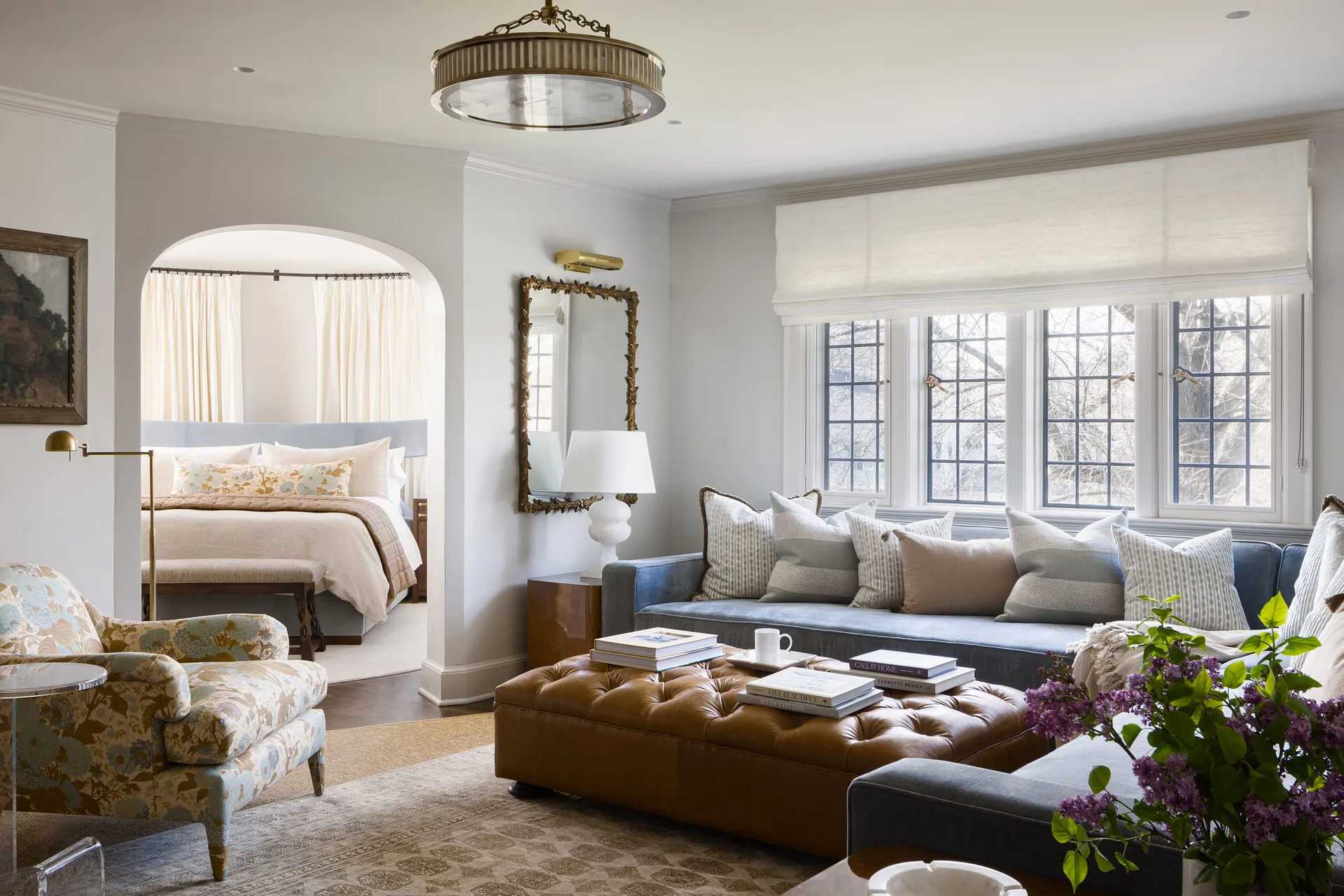 A sitting area with a blue sofa and tufted leather ottoman opens through an archway into an adjacent bedroom.