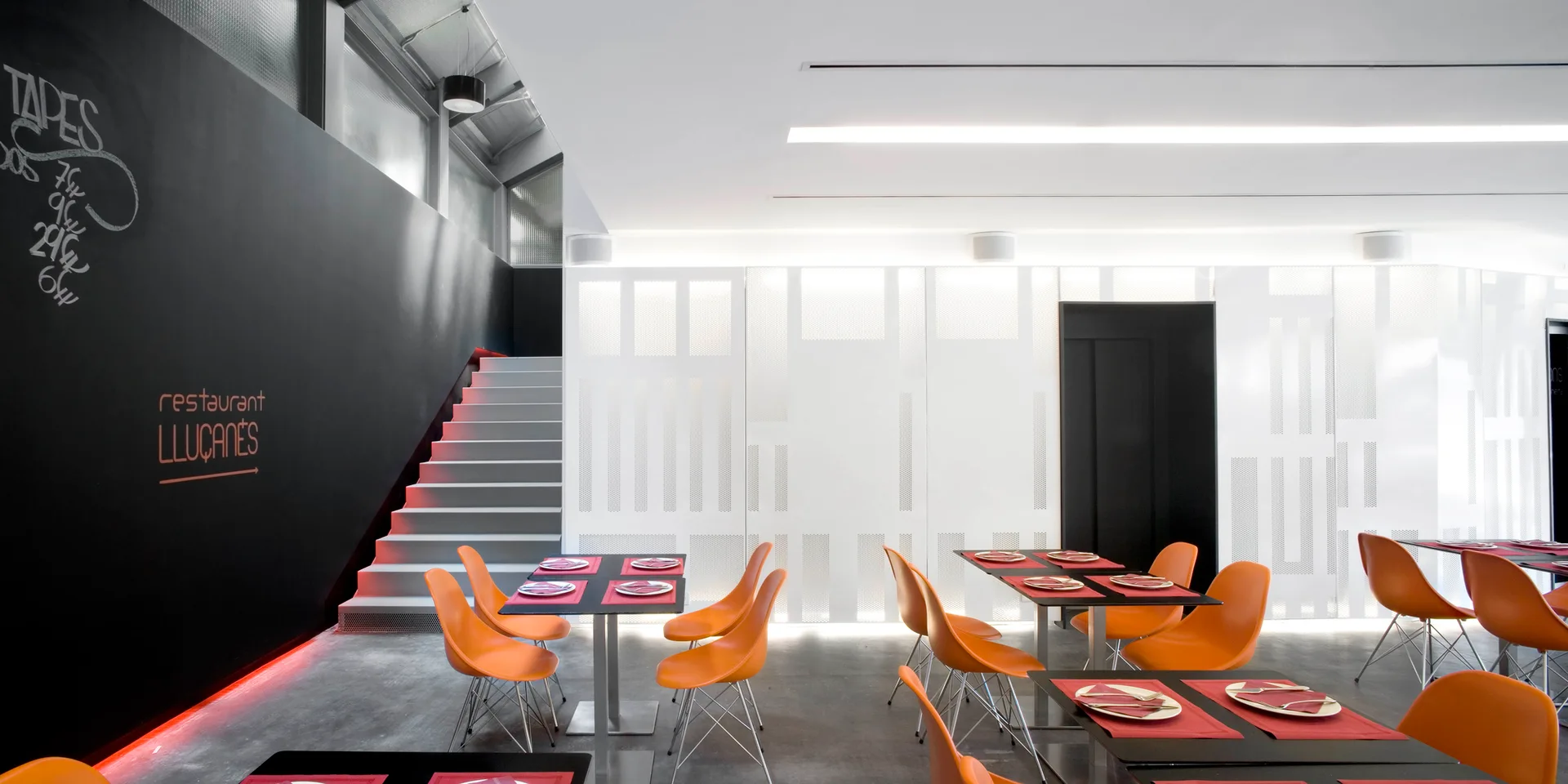 Orange chairs are arranged around dark tables in a modern dining room featuring a black wall, white staircase, and perforated white panels.