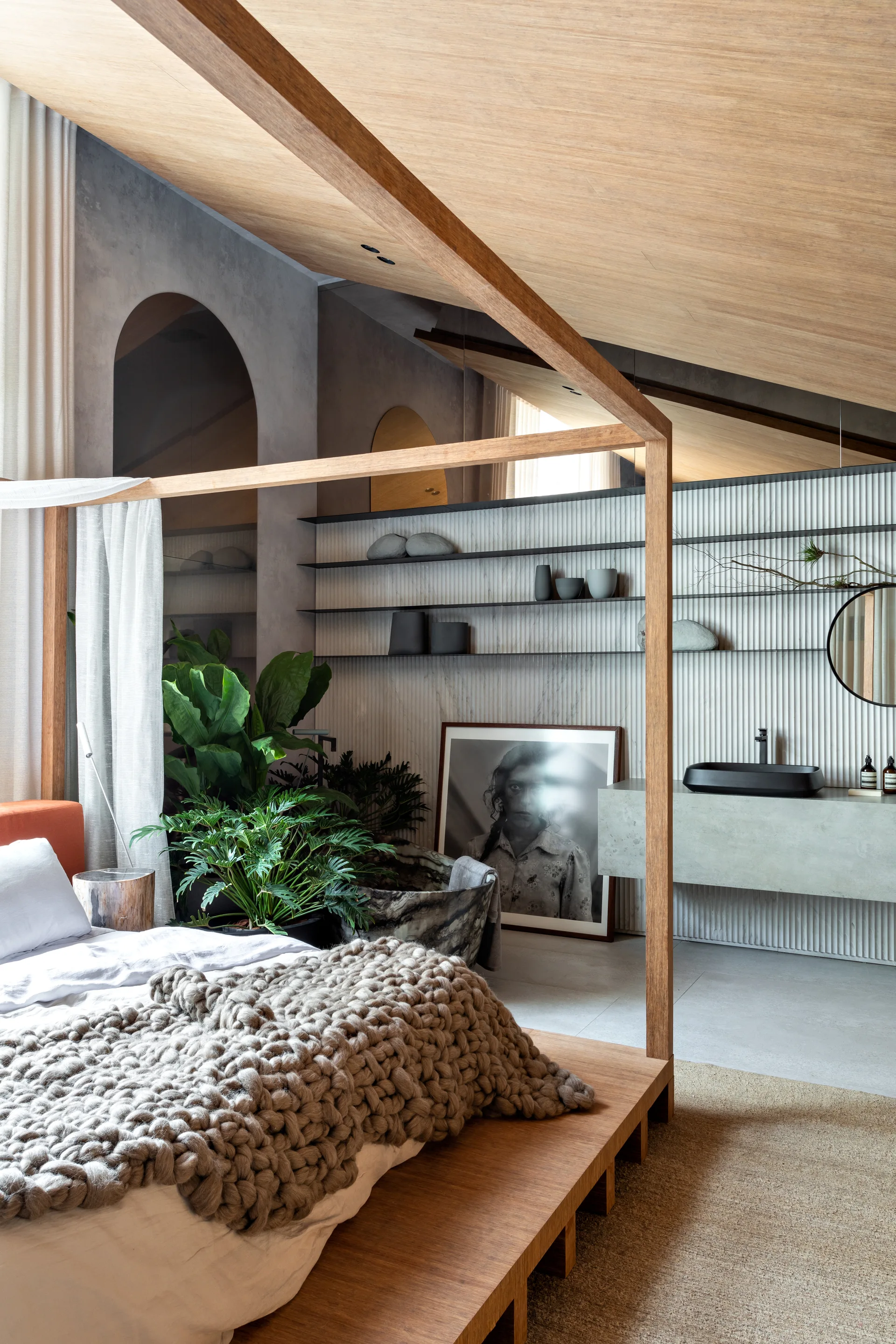 An open-plan bedroom features a natural wood four-poster bed, a chunky knit throw, and an integrated bathroom vanity against a white fluted wall.