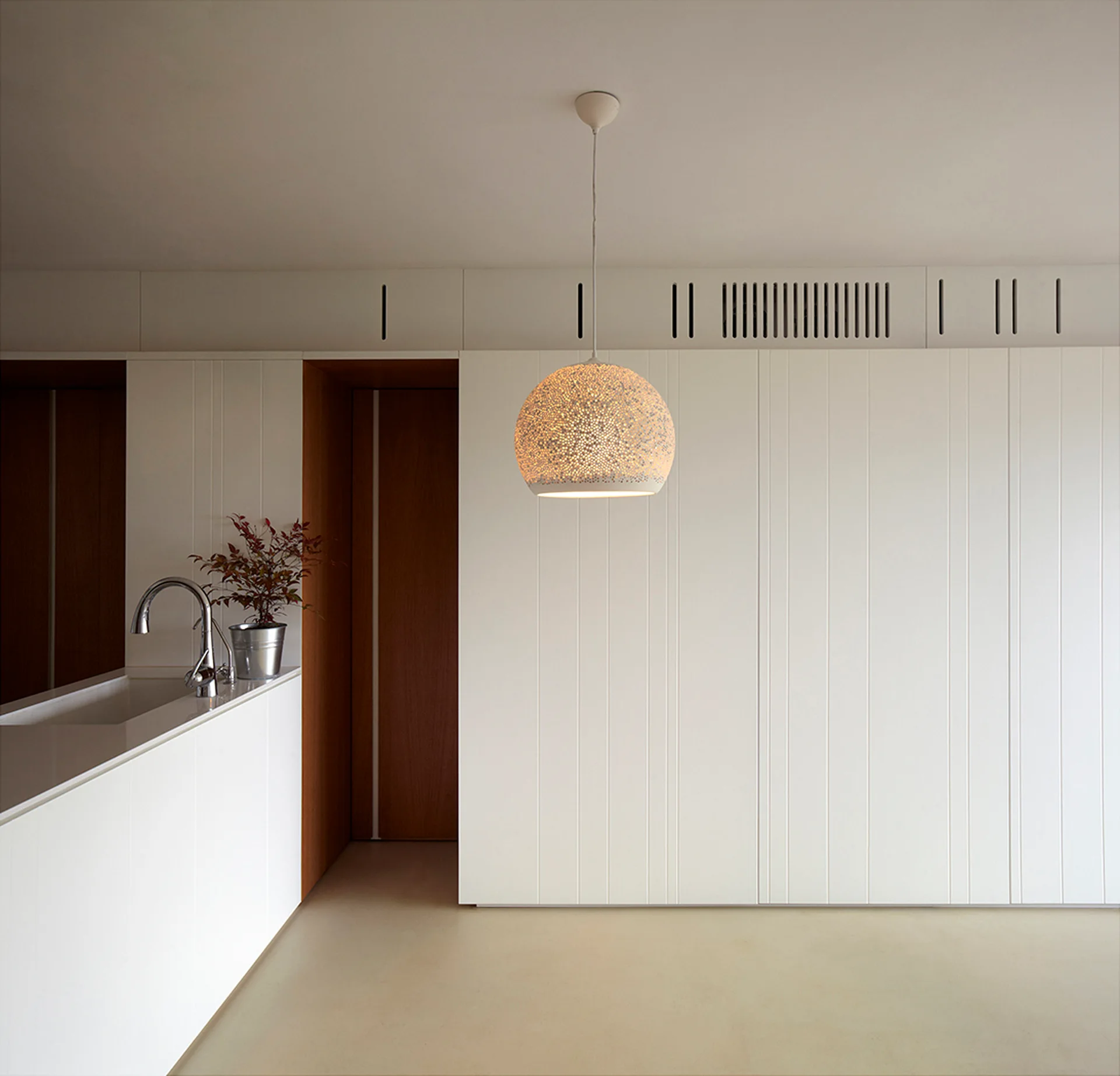 A minimalist kitchen features white vertically paneled cabinets, a wood-lined doorway, and a glowing perforated pendant light.