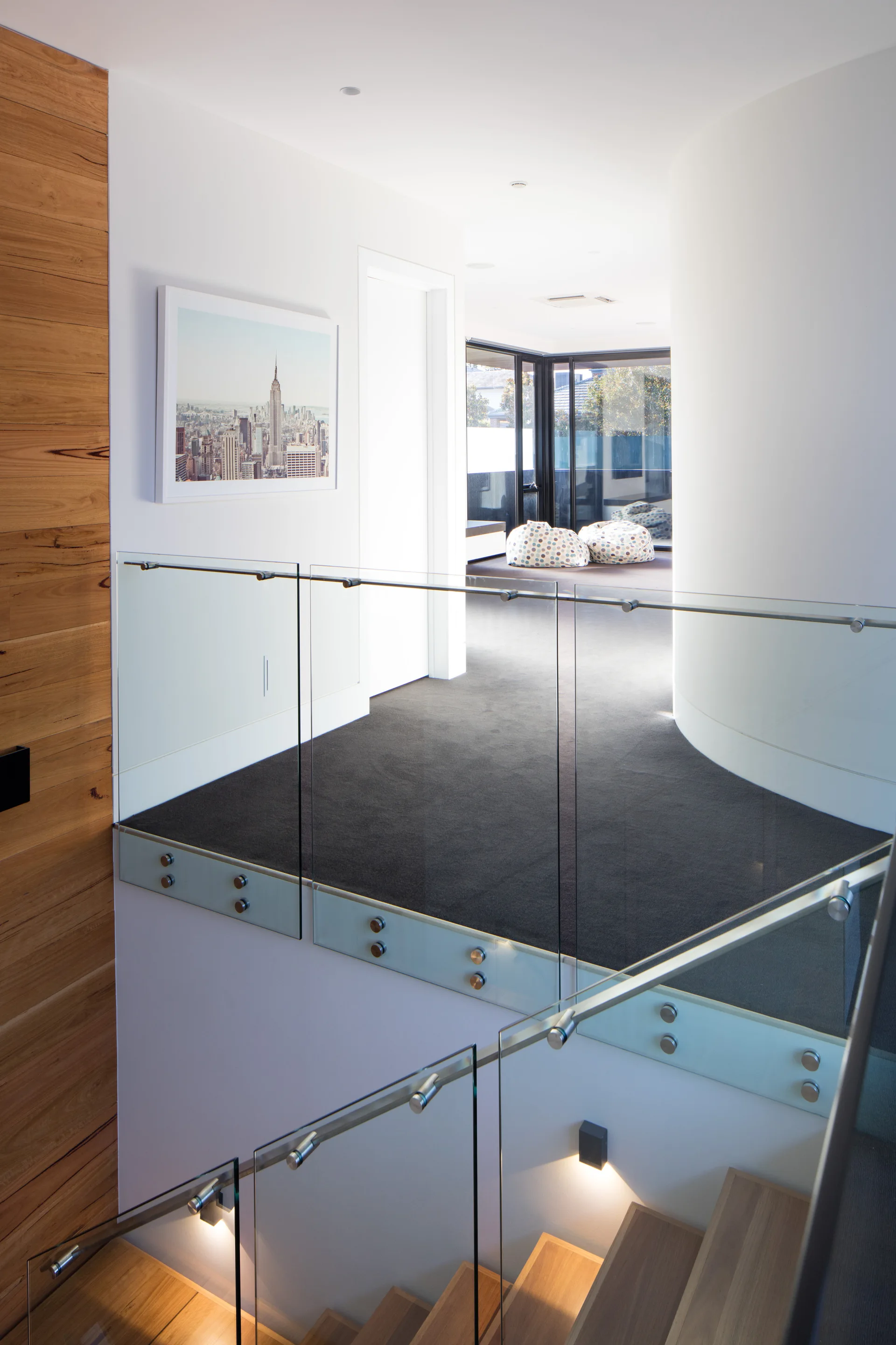 A modern wooden staircase with clear glass guardrails leads to a landing featuring dark gray carpeting, a curved white wall, and recessed lighting.