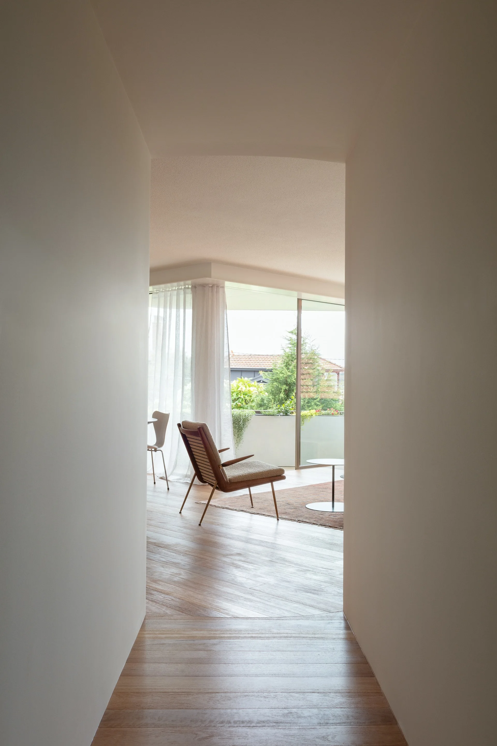 A view through a narrow off-white hallway looking into a bright living room with hardwood floors, a mid-century armchair, and sheer curtains.