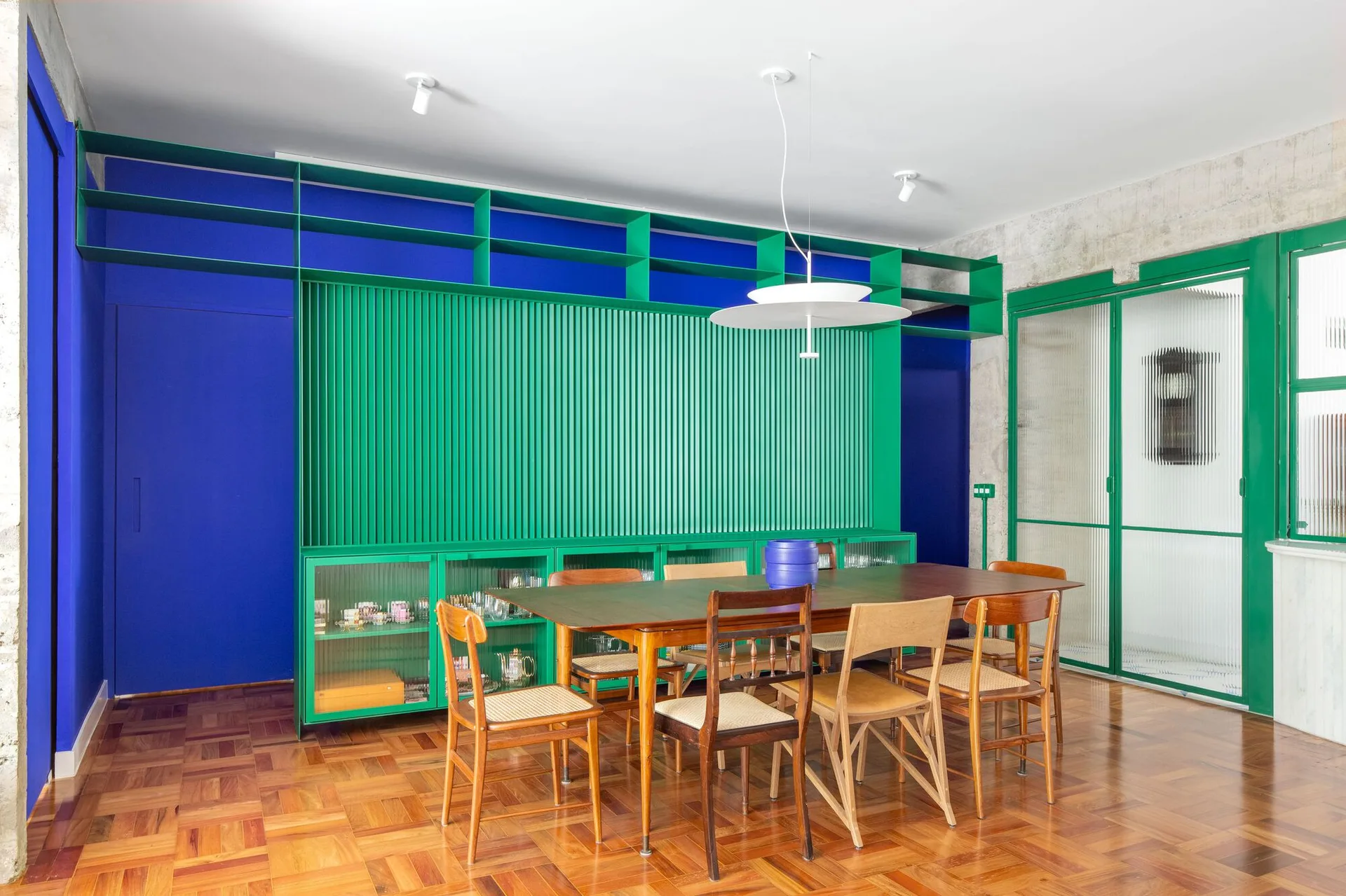 A dark wood dining table and mismatched chairs sit in a room with a large green slatted cabinet wall and a cobalt blue accent wall.
