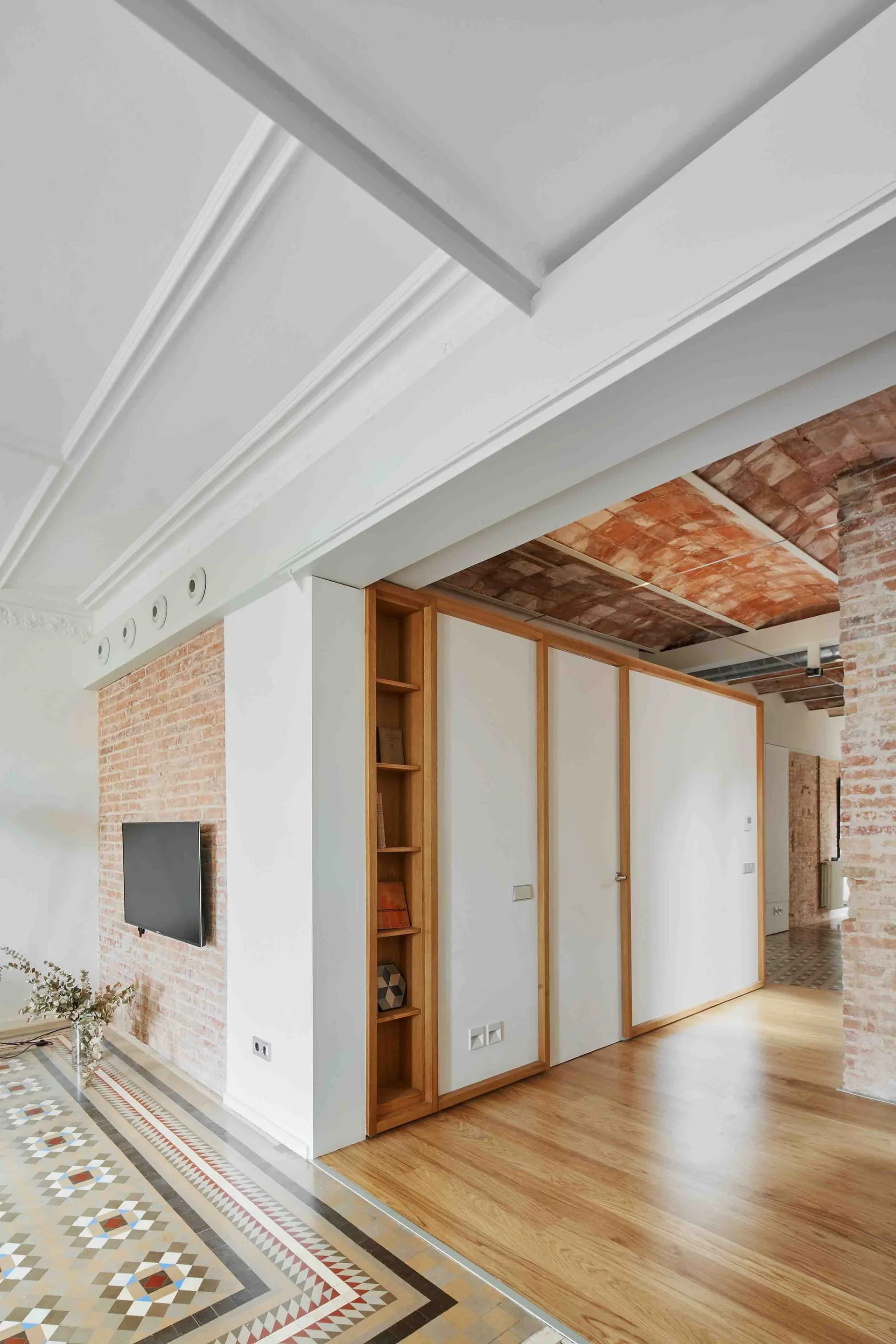 An interior space showing a transition from geometric floor tiles to wood planks, alongside exposed brick walls and a large built-in storage cabinet.