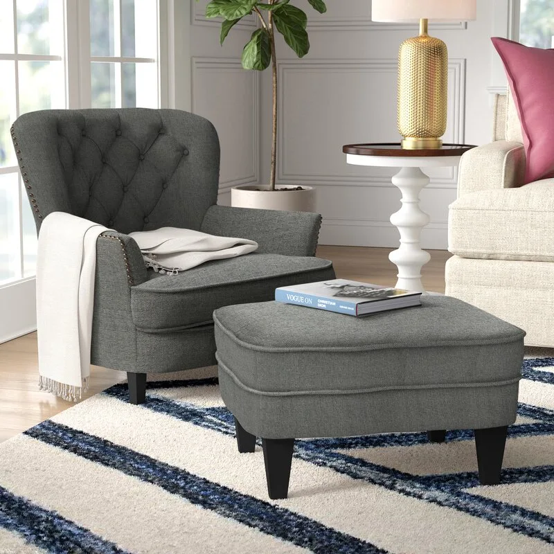A grey tufted armchair with a white throw blanket sits next to a matching ottoman on a striped blue and white rug.