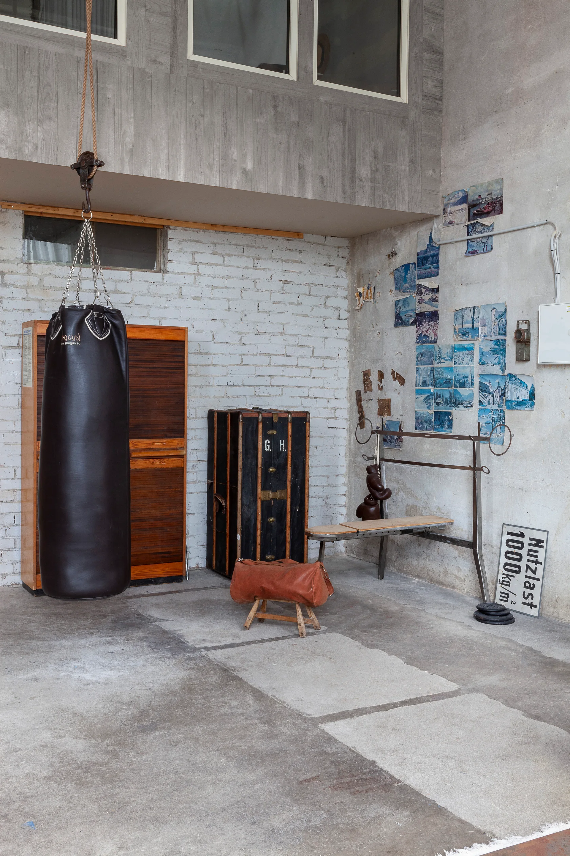 A vintage gym room features a black punching bag hanging next to a wooden cabinet, a black trunk, and a workout bench against a painted white brick wall.