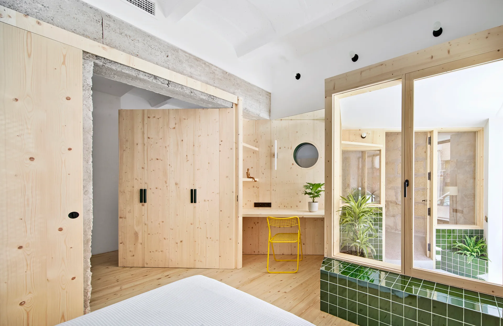 A modern bedroom featuring knotty pine wood paneling, a built-in desk with a yellow chair, and a glass partition with a green tiled base.