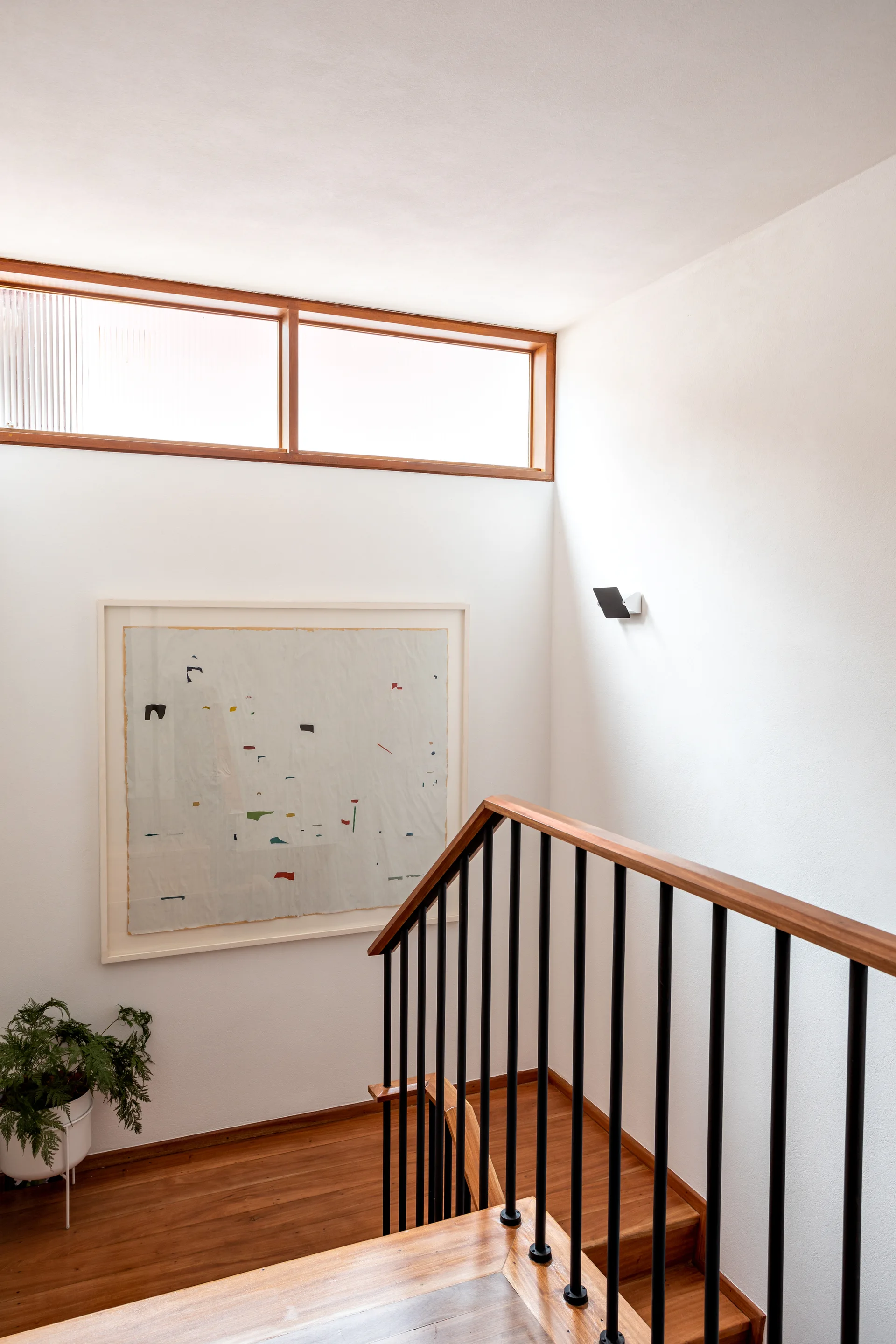 A minimalist wooden staircase with black metal railing stands next to a white wall featuring a large abstract art piece and a high transom window.