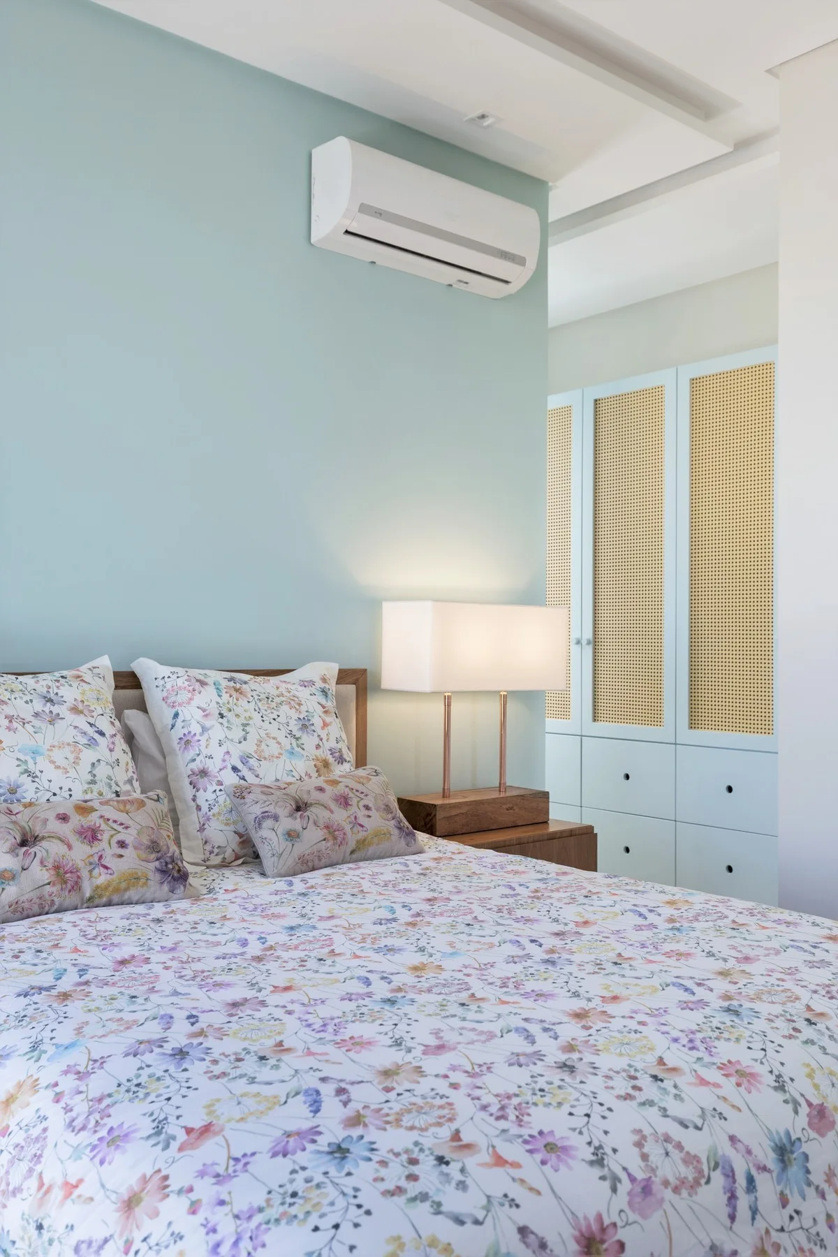 A light blue bedroom featuring a bed with floral bedding, a wood nightstand with a lamp, and a built-in wardrobe with cane door panels.