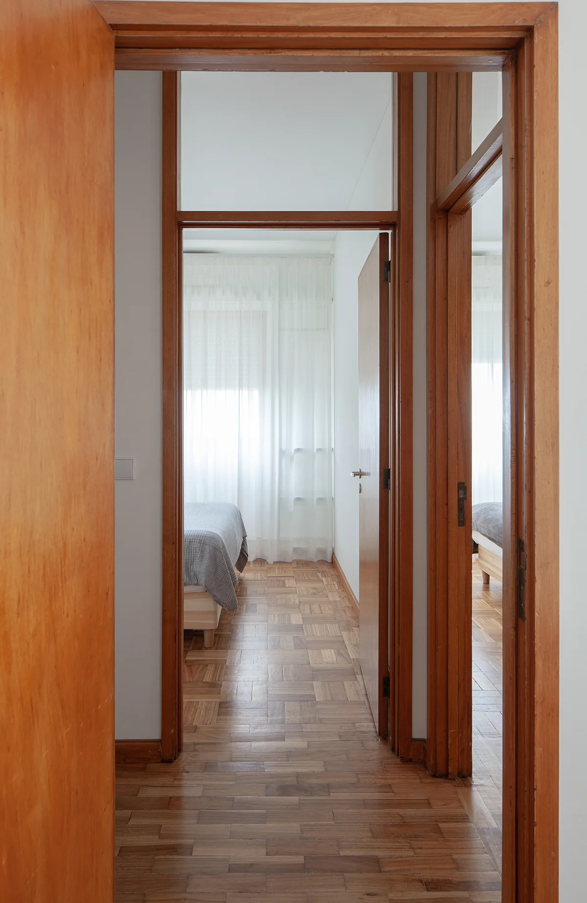 A narrow hallway with wooden parquet flooring and warm wood door frames leads to a bright bedroom with a bed and white sheer curtains.