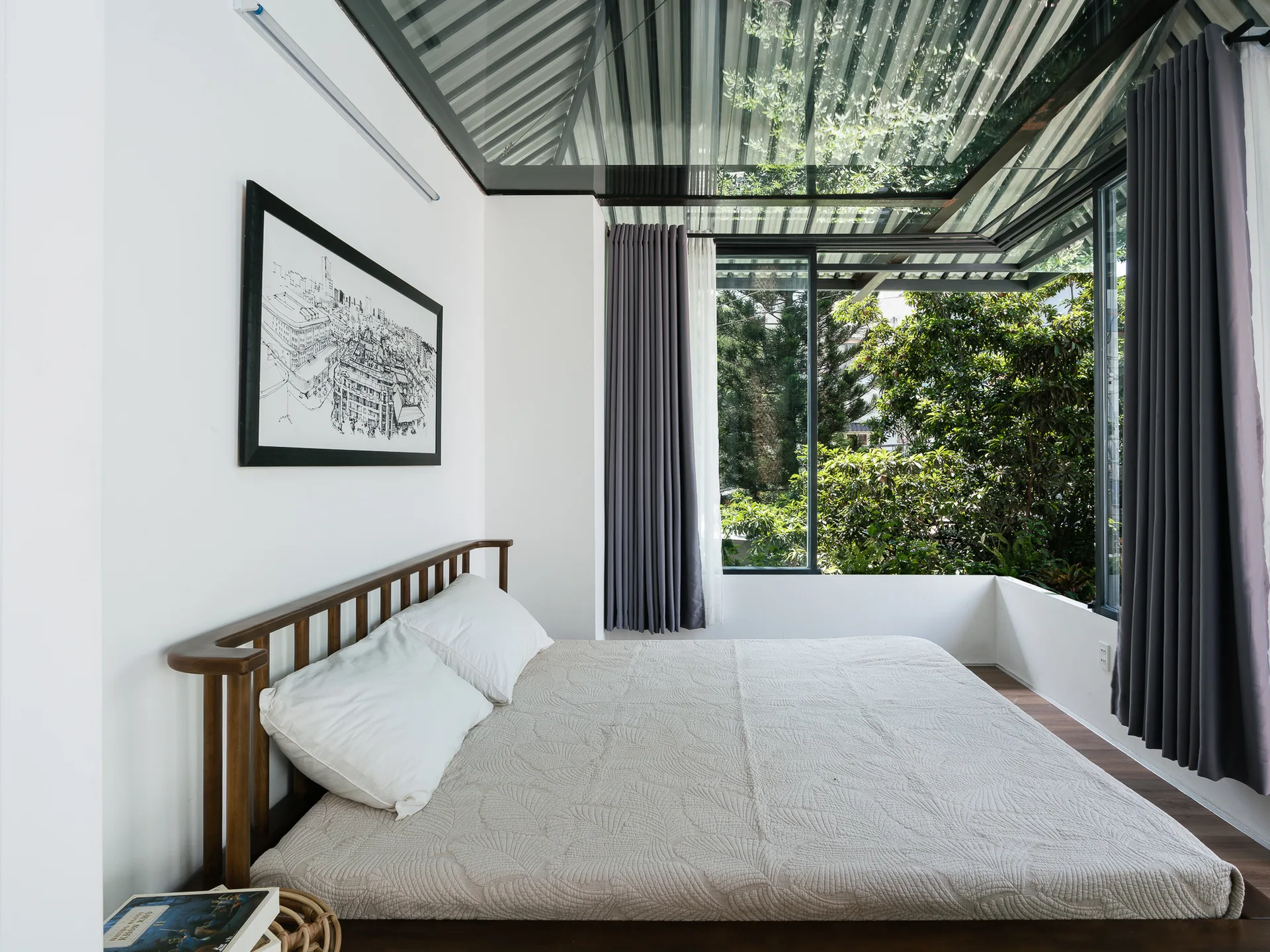 A modern bedroom features a wooden bed frame with white pillows, a large window overlooking green trees, and a framed sketch on a white wall.