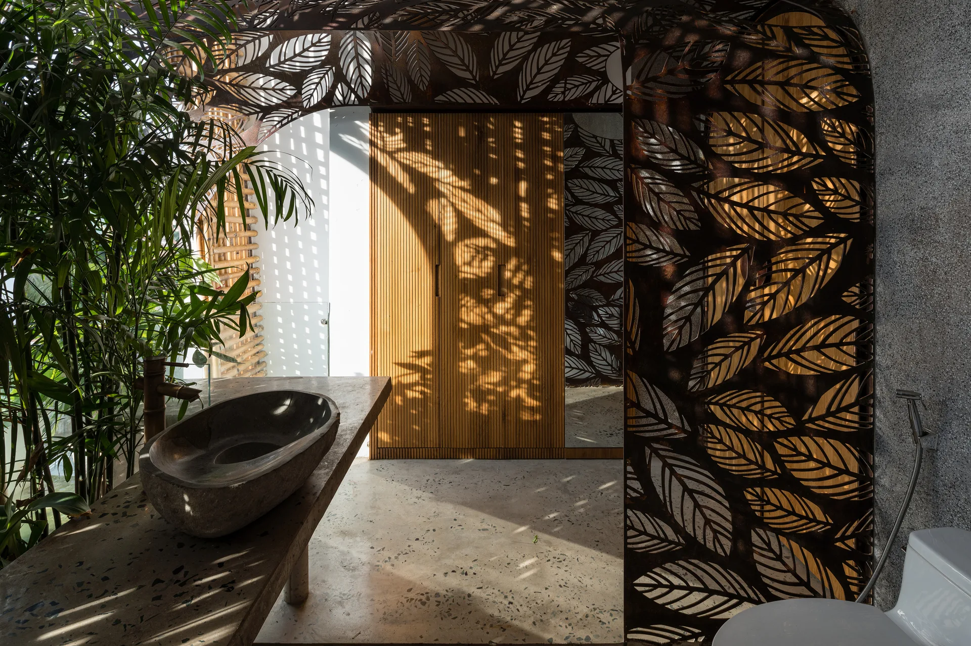 A modern bathroom with a dark stone vessel sink, a slatted wood cabinet, and a laser-cut metal screen casting leaf-shaped shadows.