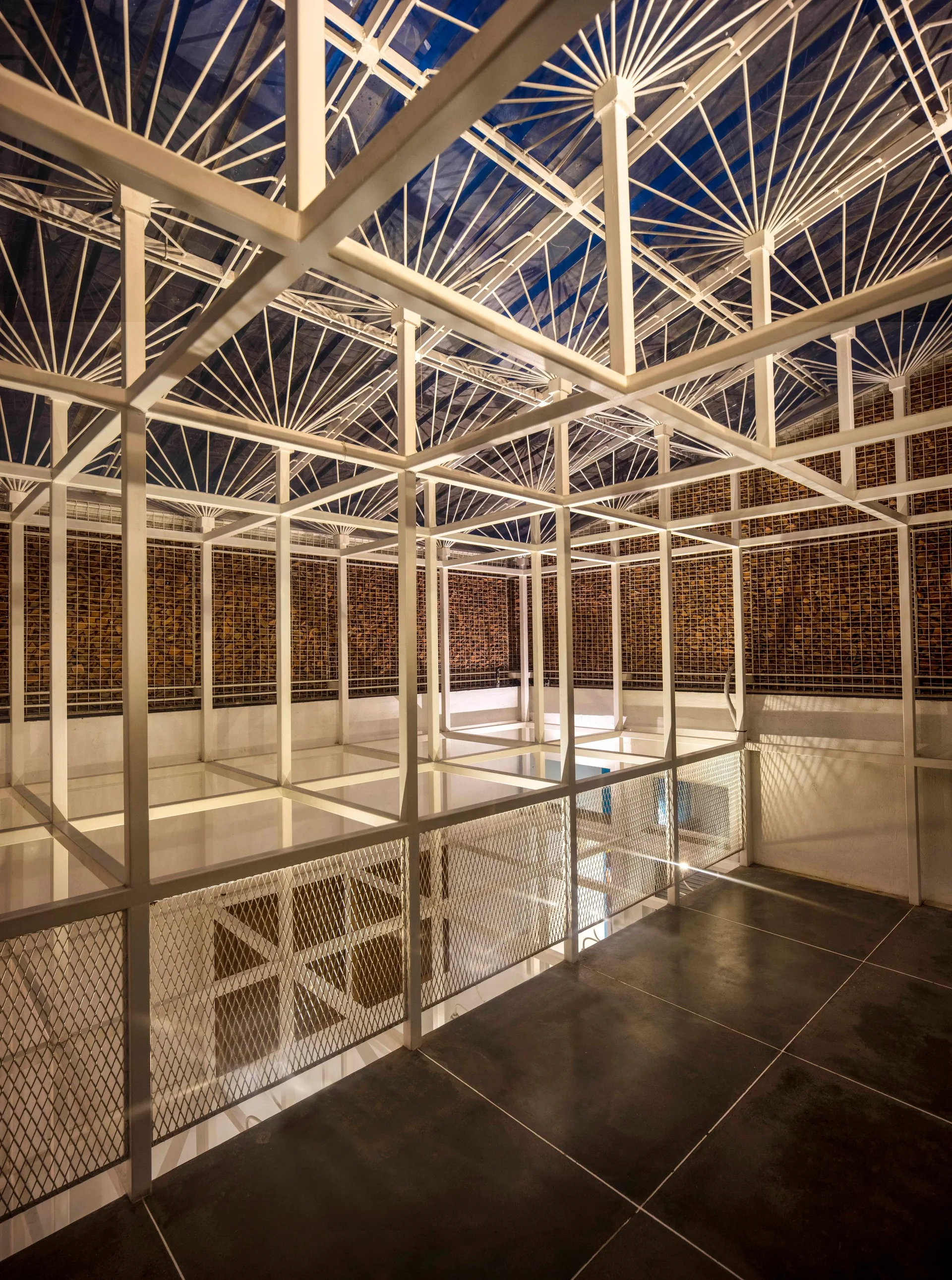 A mezzanine interior featuring a white geometric metal grid ceiling, wire mesh railings, and a wall of stacked wood behind a metal screen.