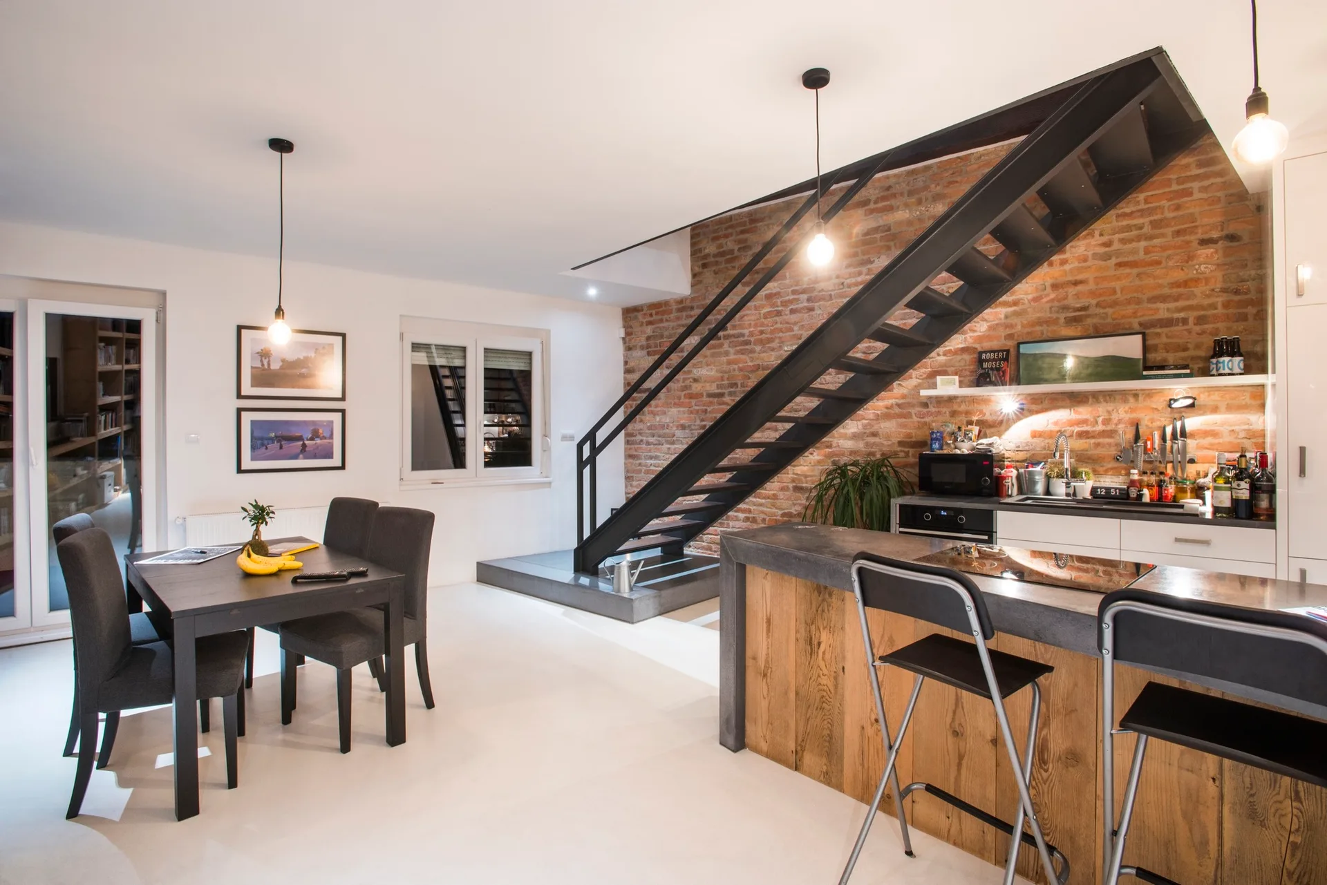An open-plan kitchen and dining area featuring a black metal staircase against an exposed brick wall, a wood-paneled island, and a dark dining table.