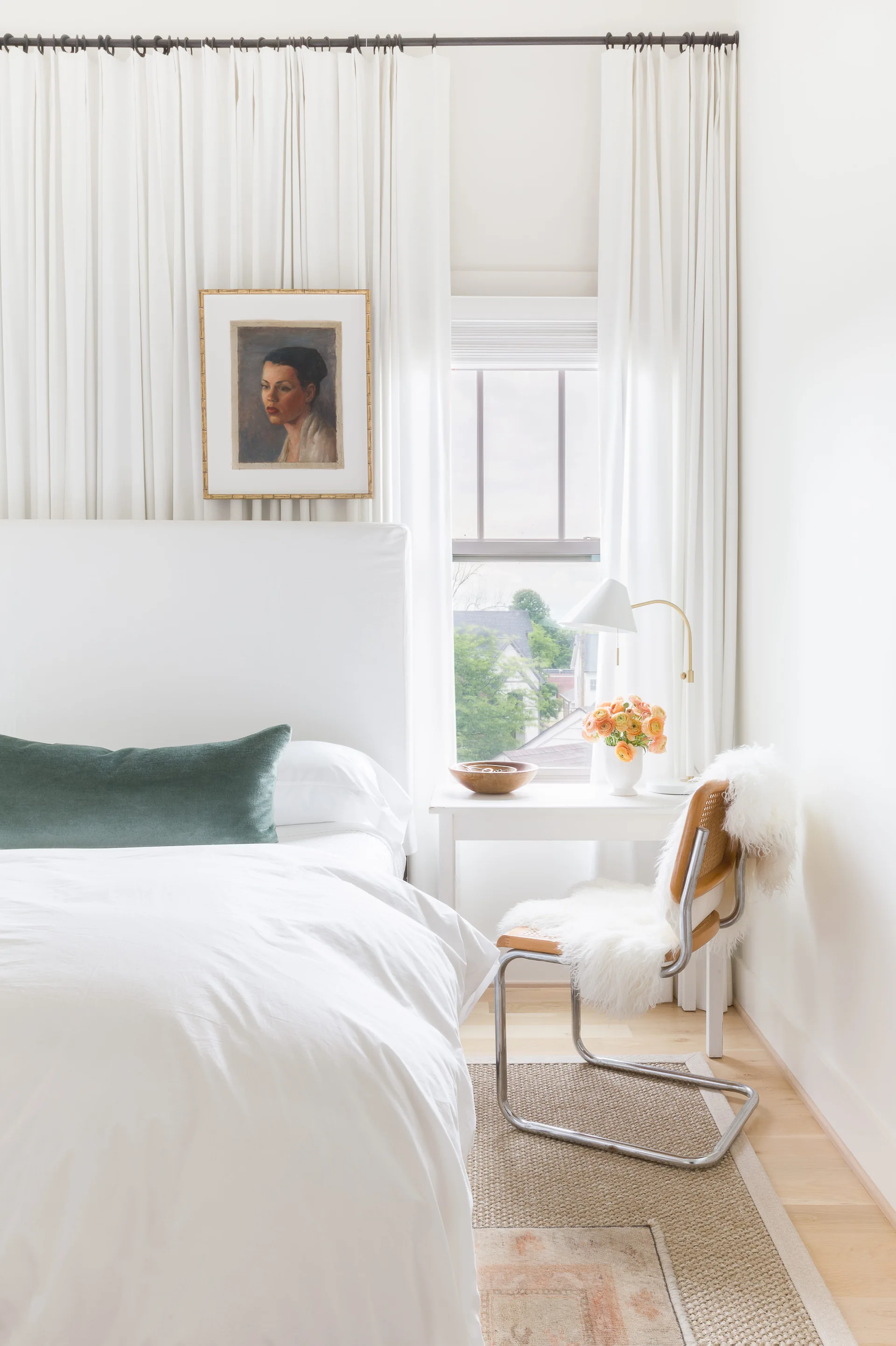 A bright white bedroom features an upholstered bed, a small white writing desk with a cane chair, and expansive floor-to-ceiling white curtains.