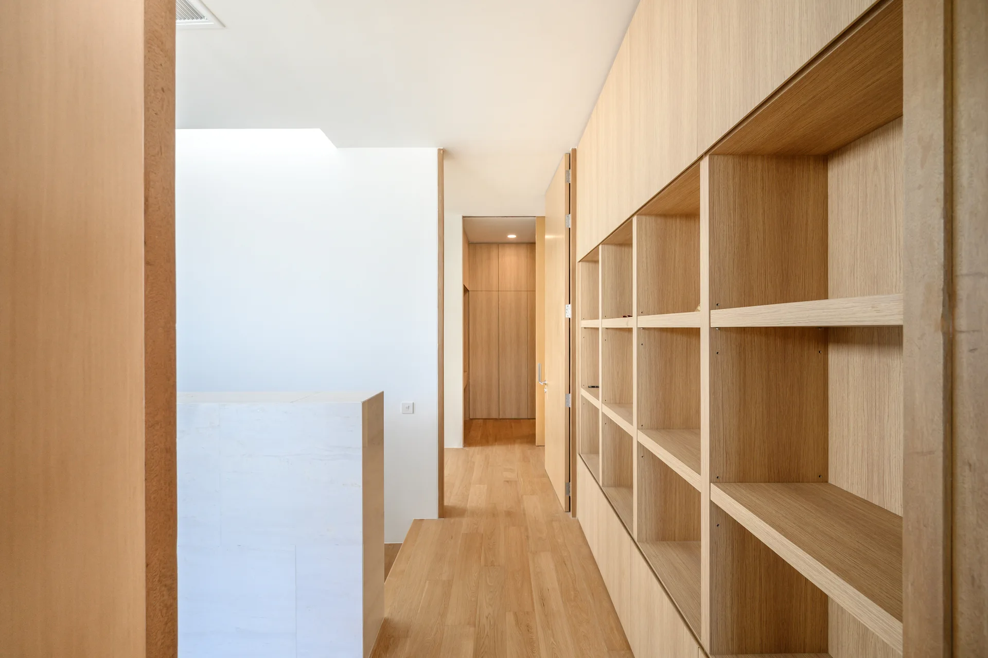 A minimalist hallway featuring a large light wood built-in shelving wall, matching wood flooring, a white partition, and open doorways.