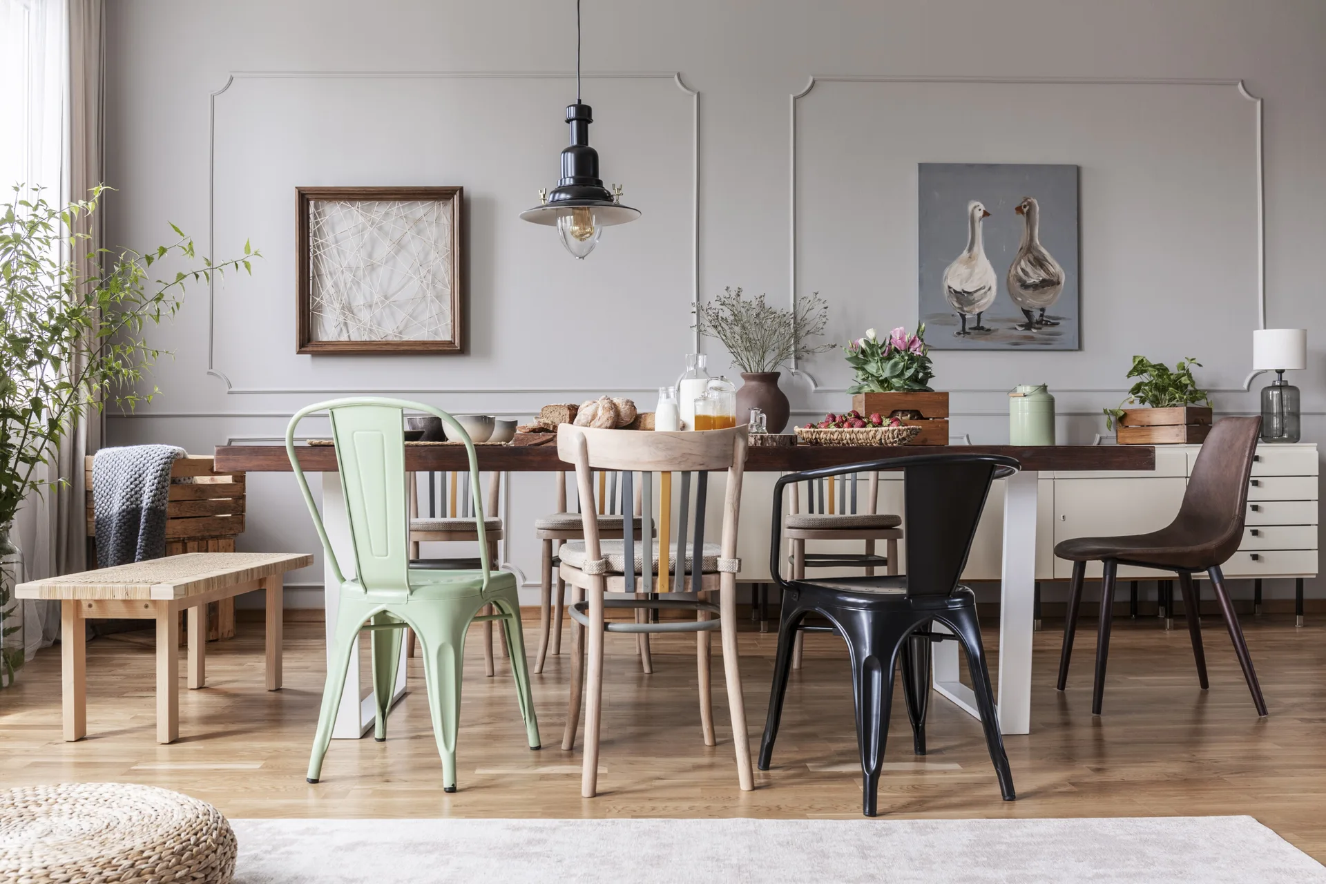 An eclectic dining room features a long wooden table set with mismatched wood and metal chairs against a grey paneled wall with a black pendant light.