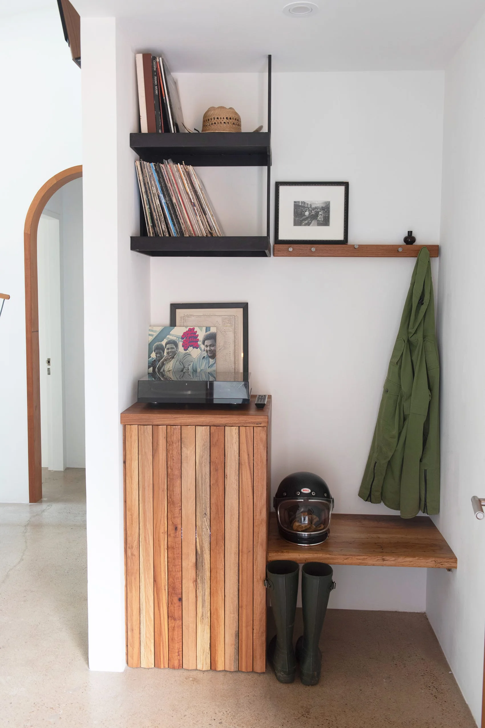 A minimalist entryway featuring a slatted wood cabinet with a record player, an integrated bench with boots, and wall-mounted shelves.