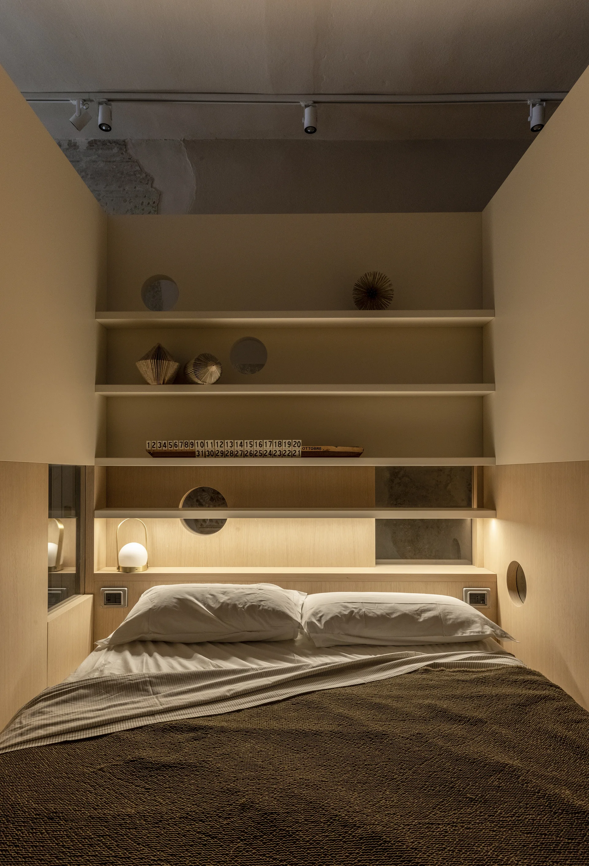 A compact bedroom alcove features a bed with white pillows and a brown blanket beneath integrated light wood shelving with circular cutouts.