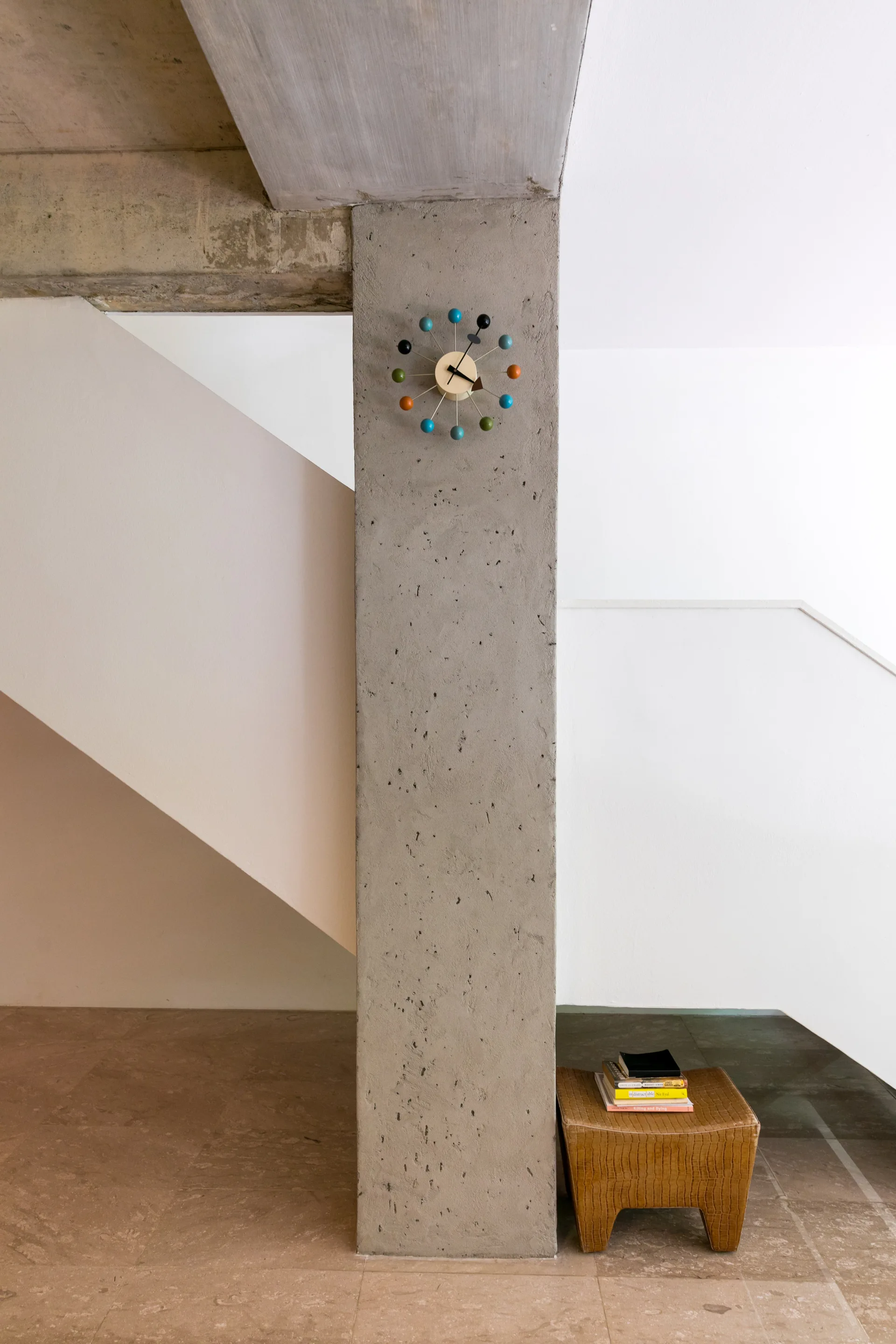 A vertical raw concrete pillar features a colorful mid-century ball clock, standing next to a white staircase and a small textured stool with books.