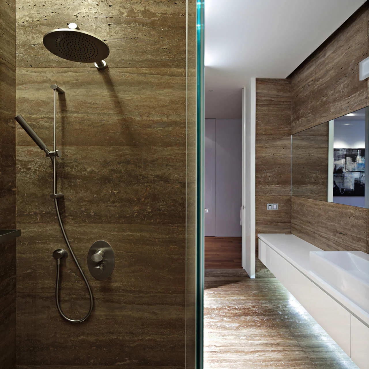 A minimalist bathroom with brown stone tile walls and floor, featuring a long white floating vanity with under-cabinet LED strip lighting.