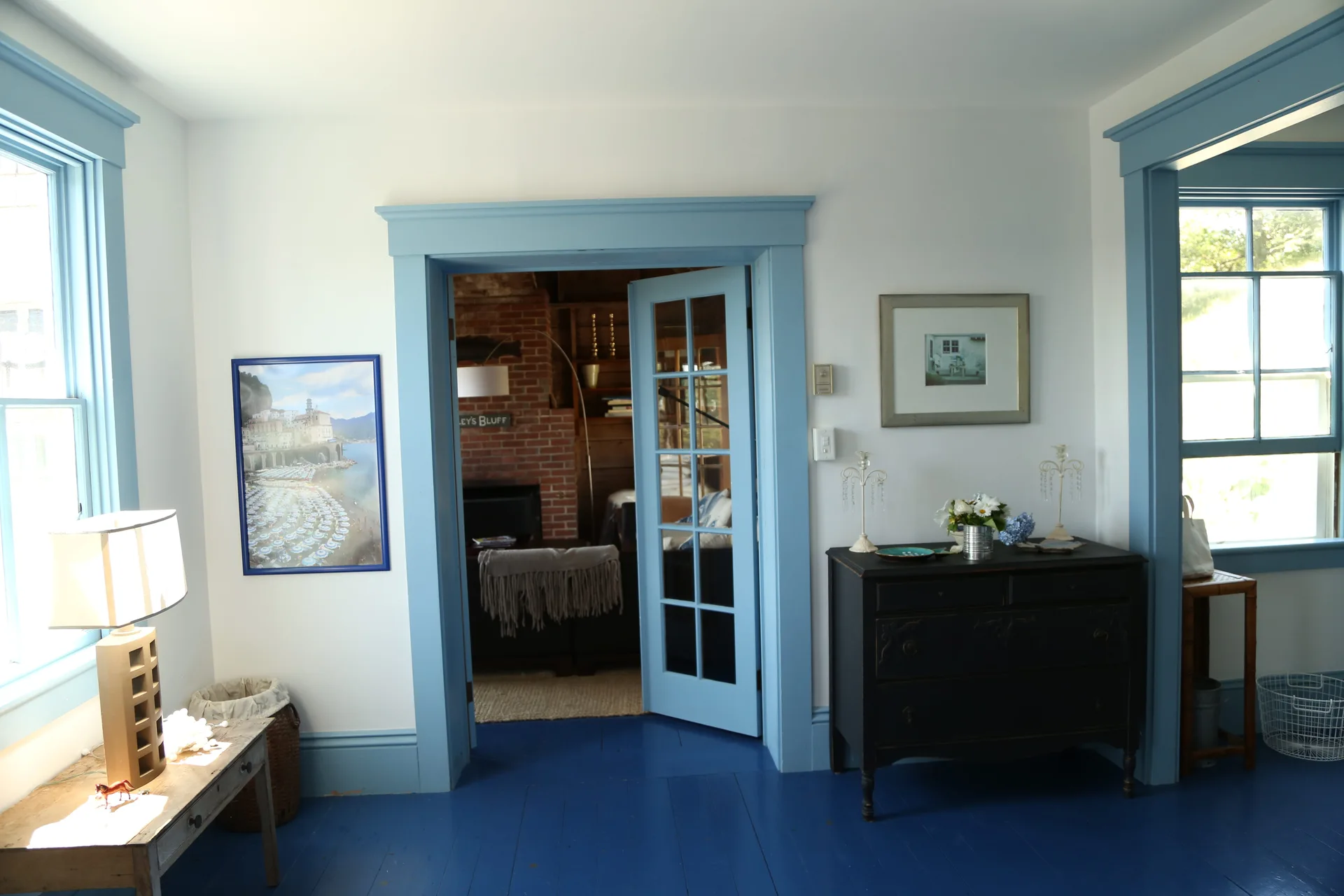 A cottage room features bright blue painted floors and matching blue trim around a French door, alongside white walls and a black wooden dresser.
